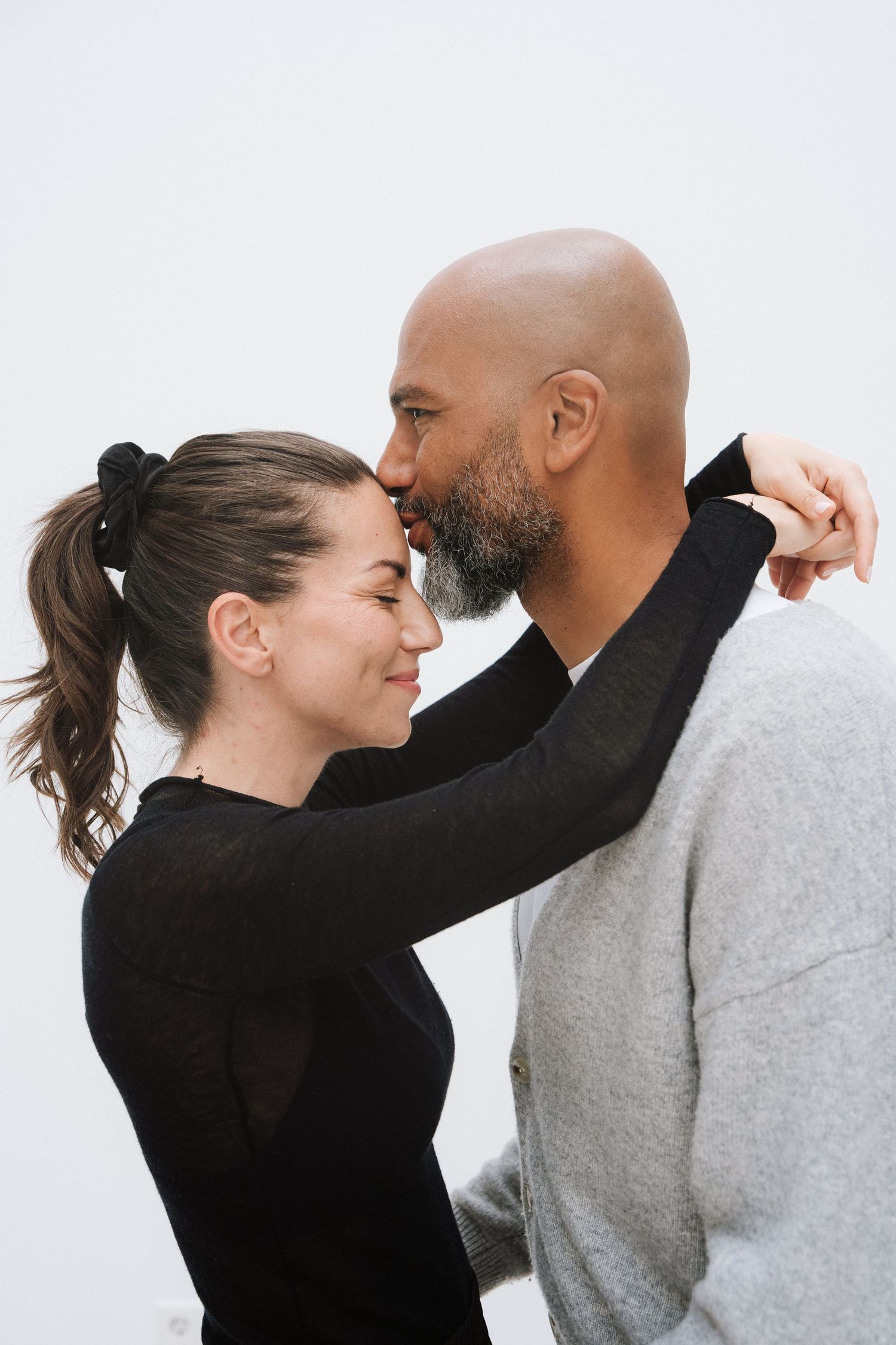 Interracial couple sharing a tender moment, man kissing woman's forehead against white background.