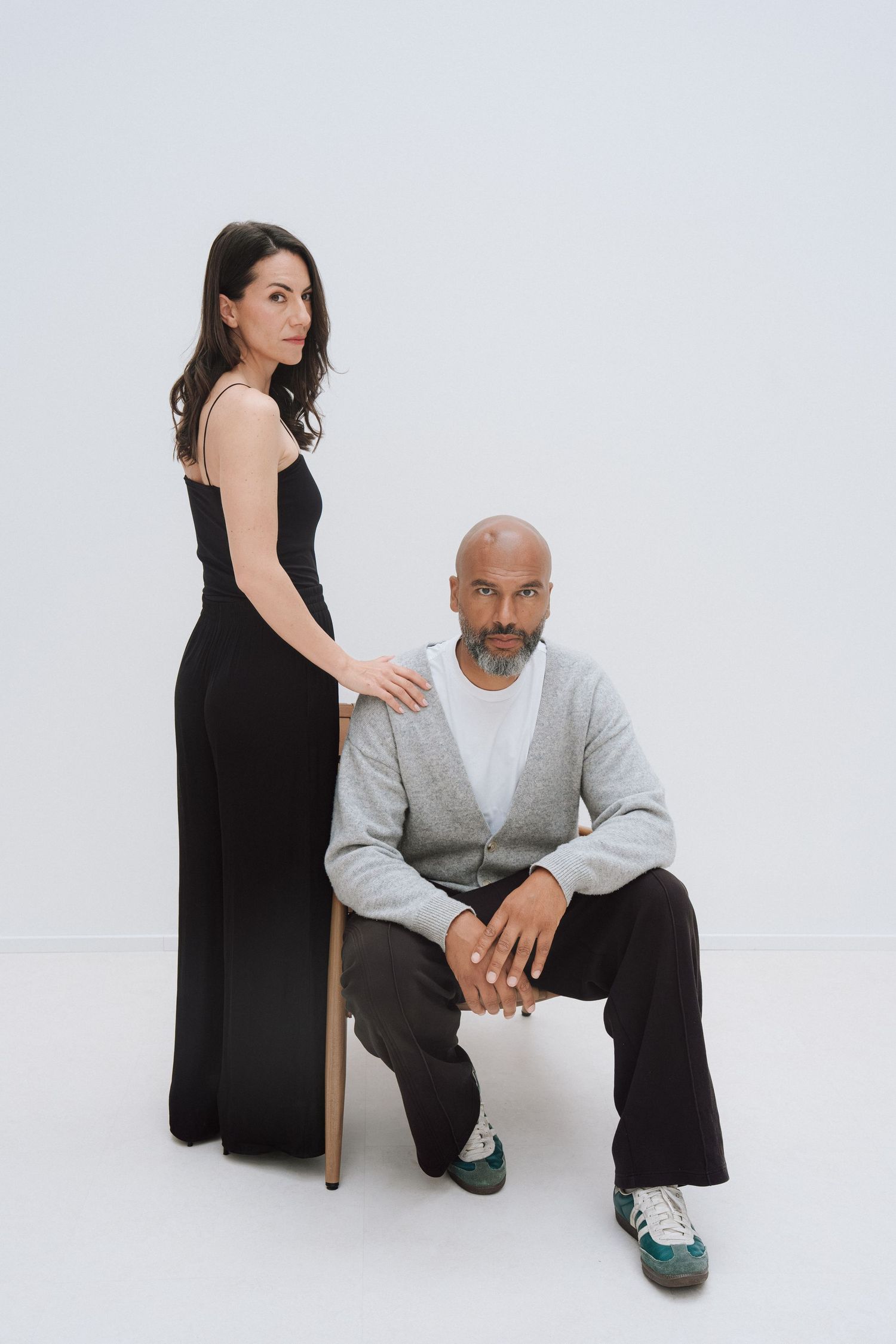 A woman in a black jumpsuit stands beside a bald bearded man seated on a chair against a white background.