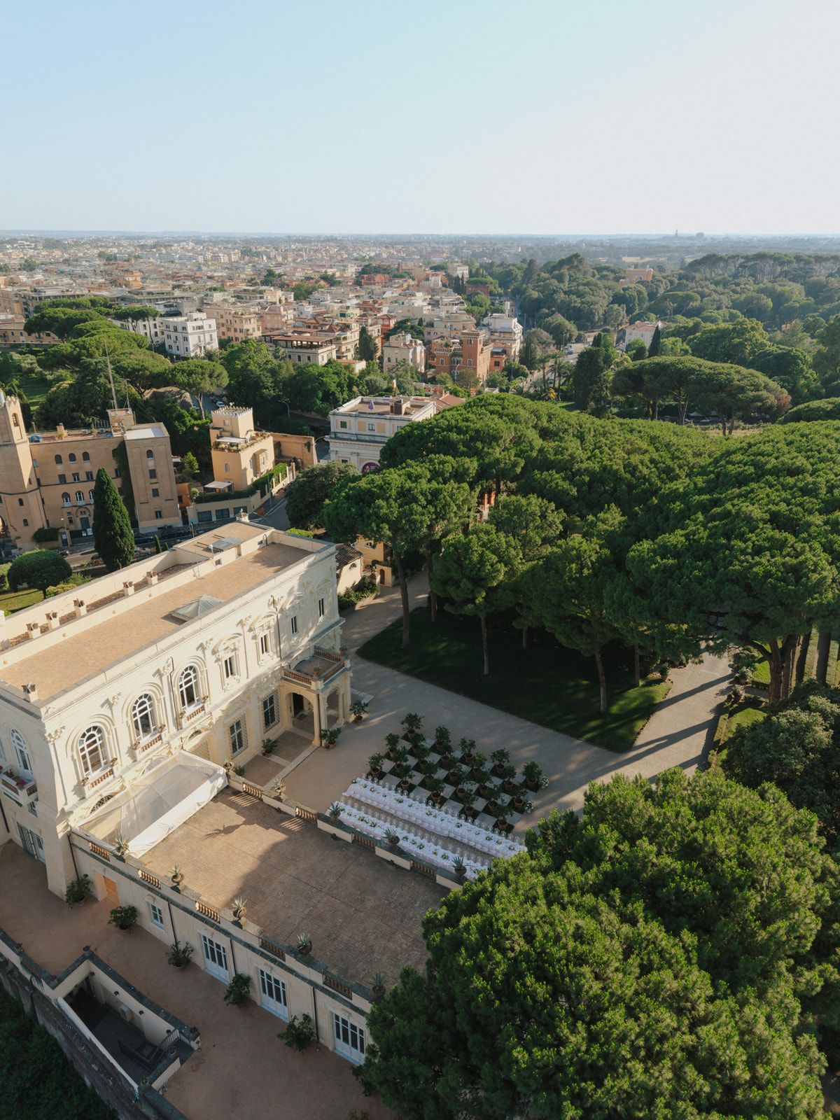 Aerial view of an elegant white villa with outdoor seating amid lush pine trees overlooking Rome cityscape.