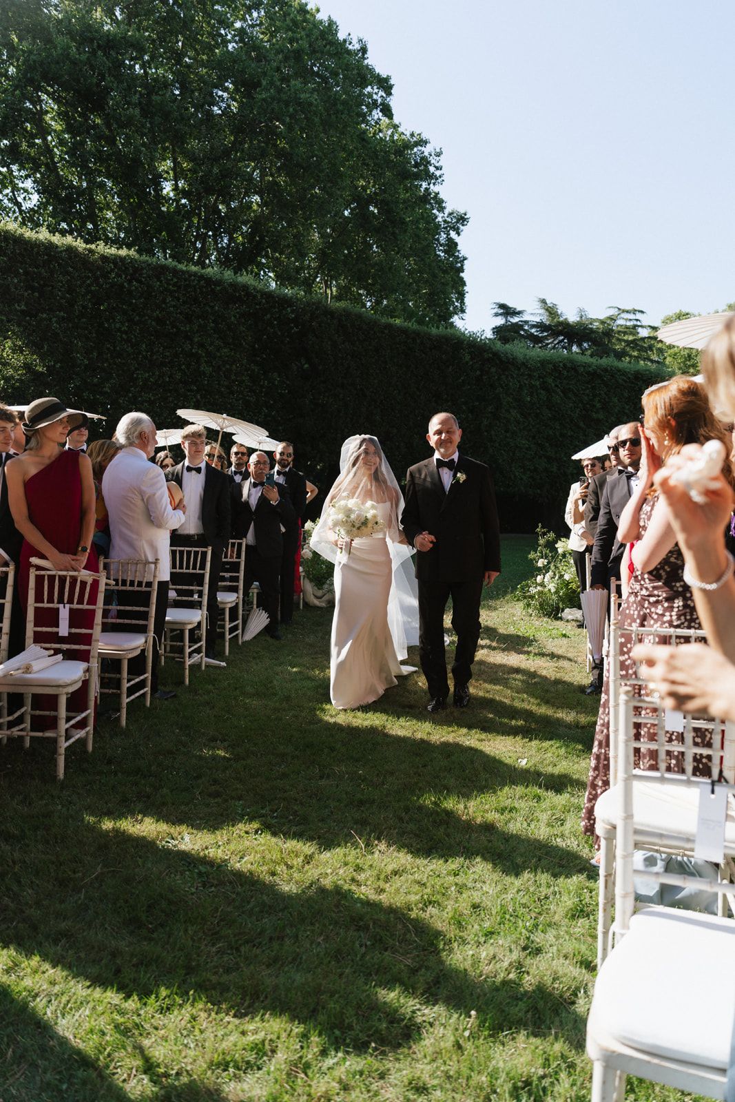 Bride walks down outdoor aisle with escort at garden wedding ceremony, flanked by guests seated on white chairs.