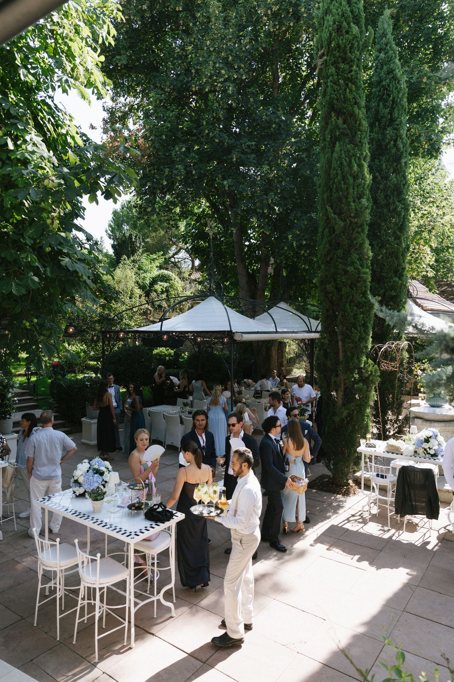 Elegant outdoor garden wedding reception with guests mingling near white floral tables under lush green trees on a sunny day.
