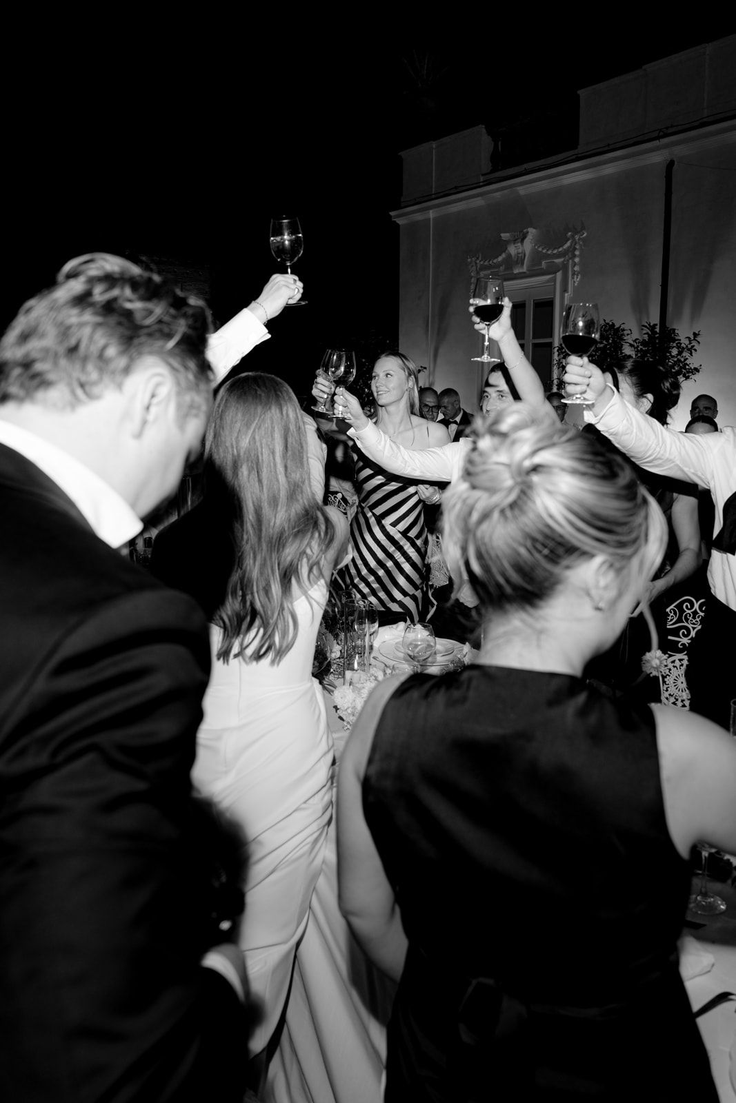 Black and white photo of wedding guests raising glasses in a celebratory toast at an elegant evening reception.
