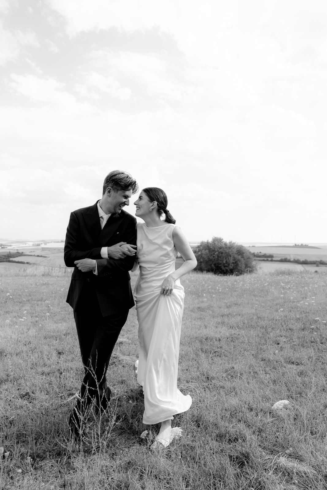 Black and white photo of a couple in wedding attire walking through a field, smiling at each other on their wedding day.