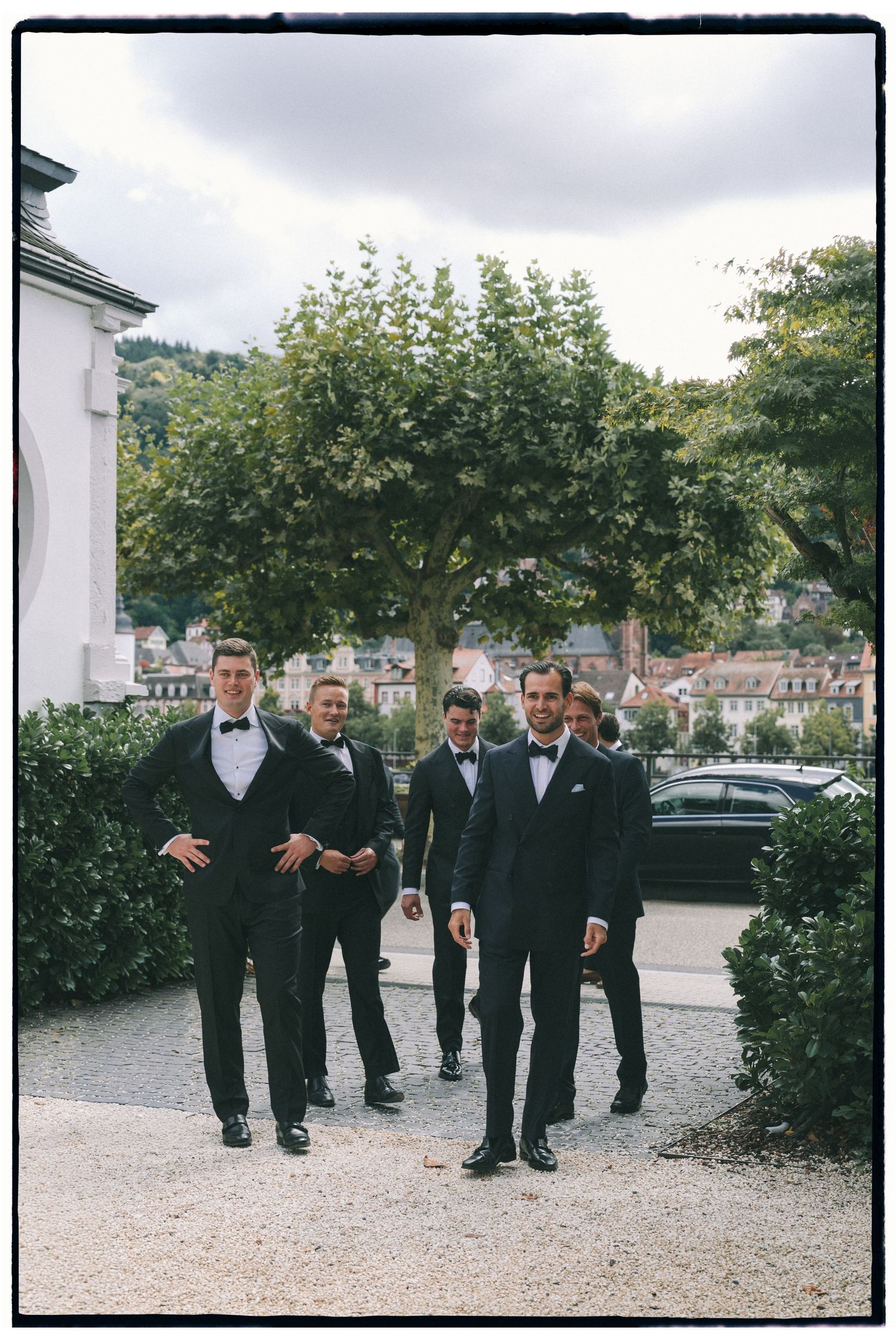 Groomsmen in black tuxedos walking together on a gravel path outdoors.