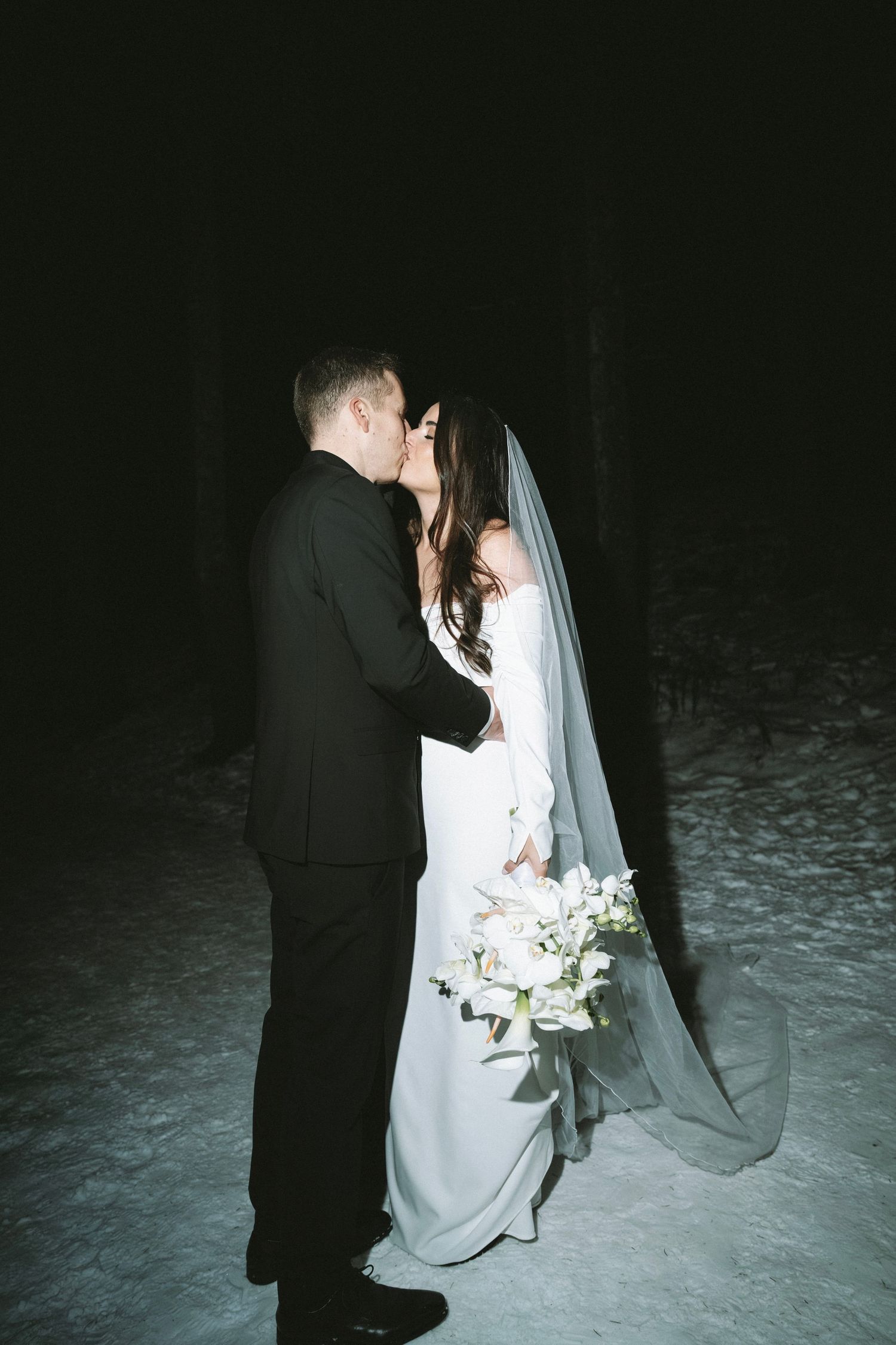 Wedding couple in formal attire taking photos in the snow at night.