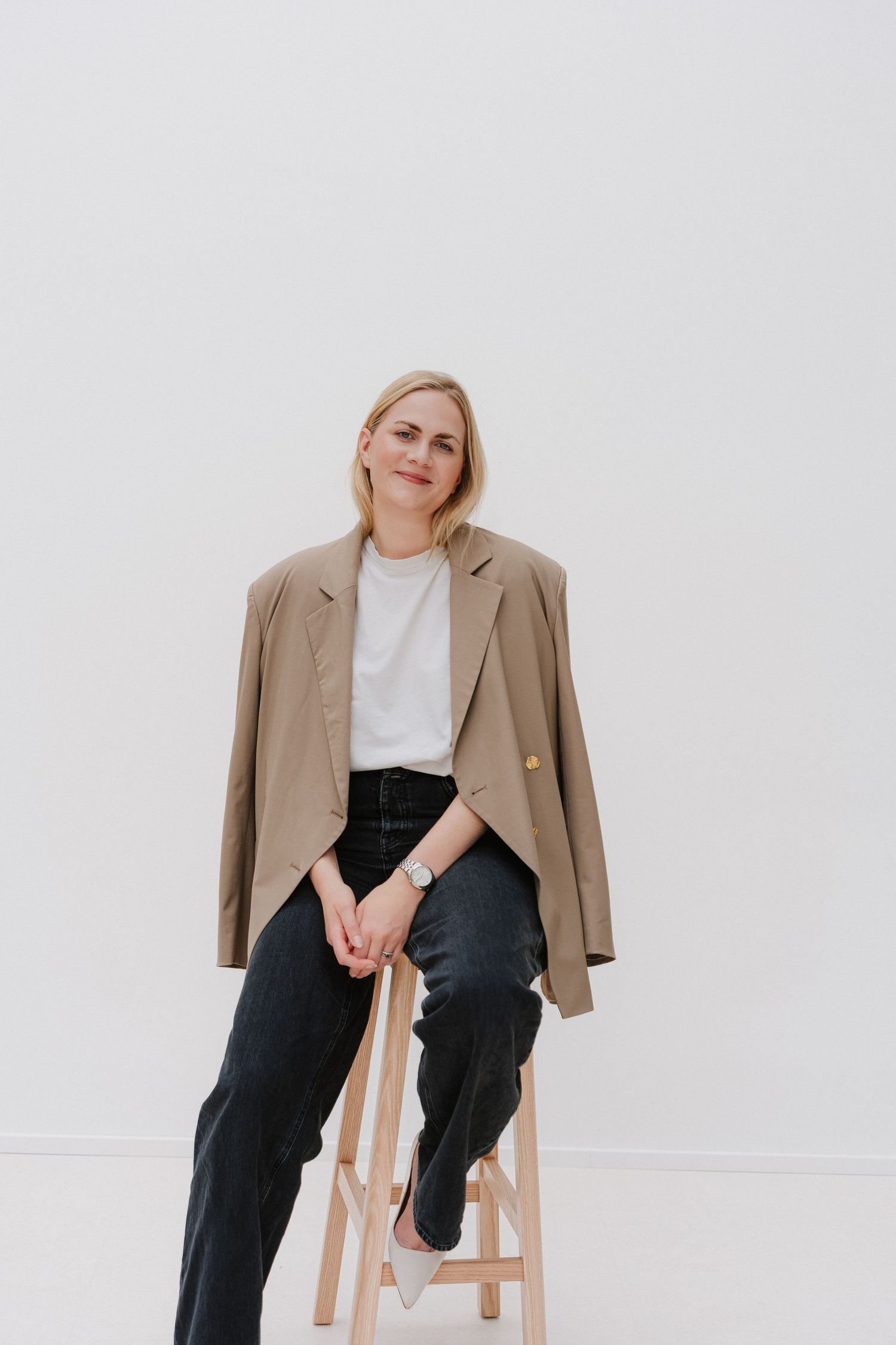 Blonde woman in beige blazer and black jeans sits on wooden stool against white background, smiling confidently.