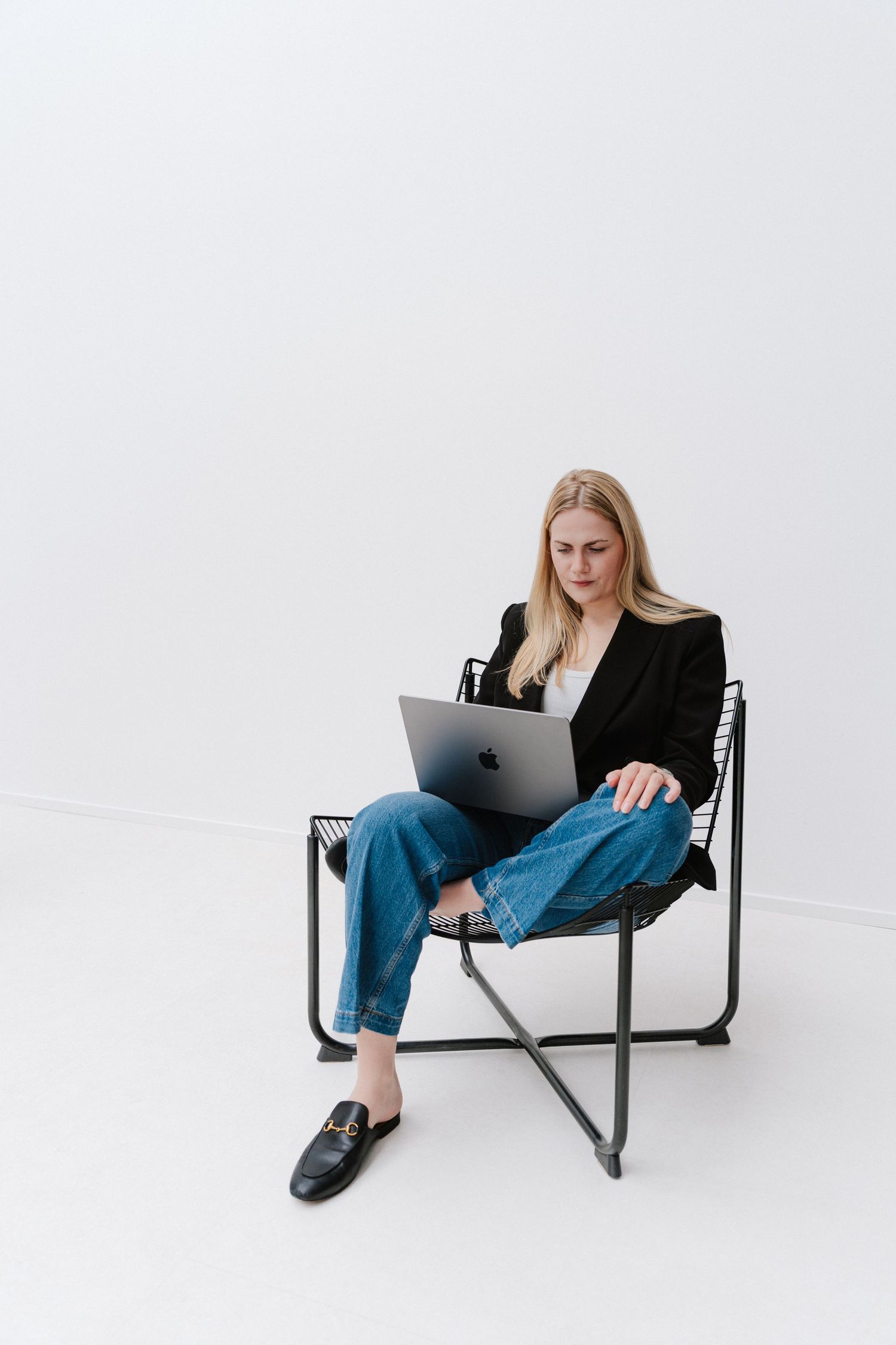 Woman in black blazer and jeans sitting in modern chair using Apple MacBook laptop on white background.