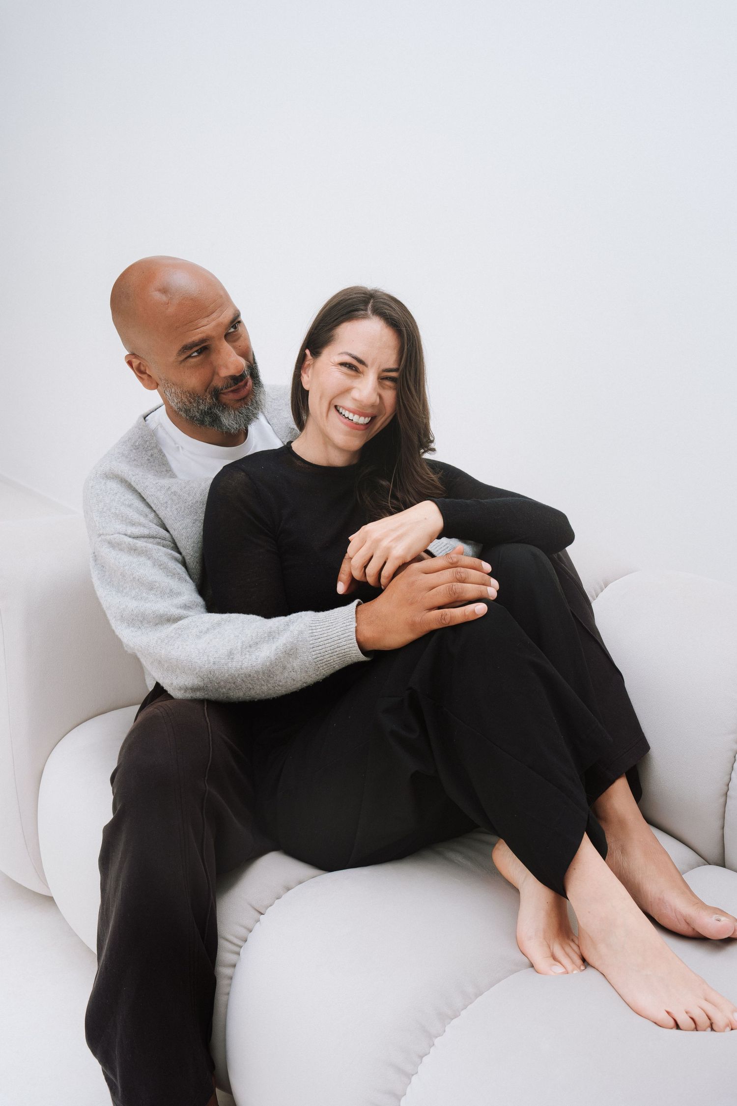 A happy couple sitting together on a white sofa, man with beard hugging smiling woman in black outfit.