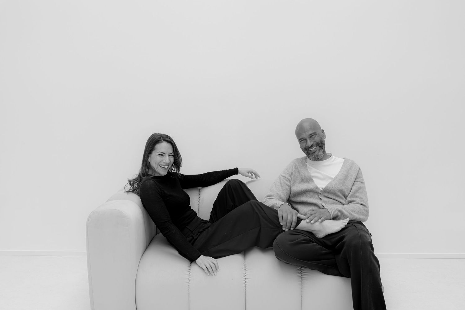 Black and white photo of a woman and bald man relaxing on a modern white sofa against a white background.