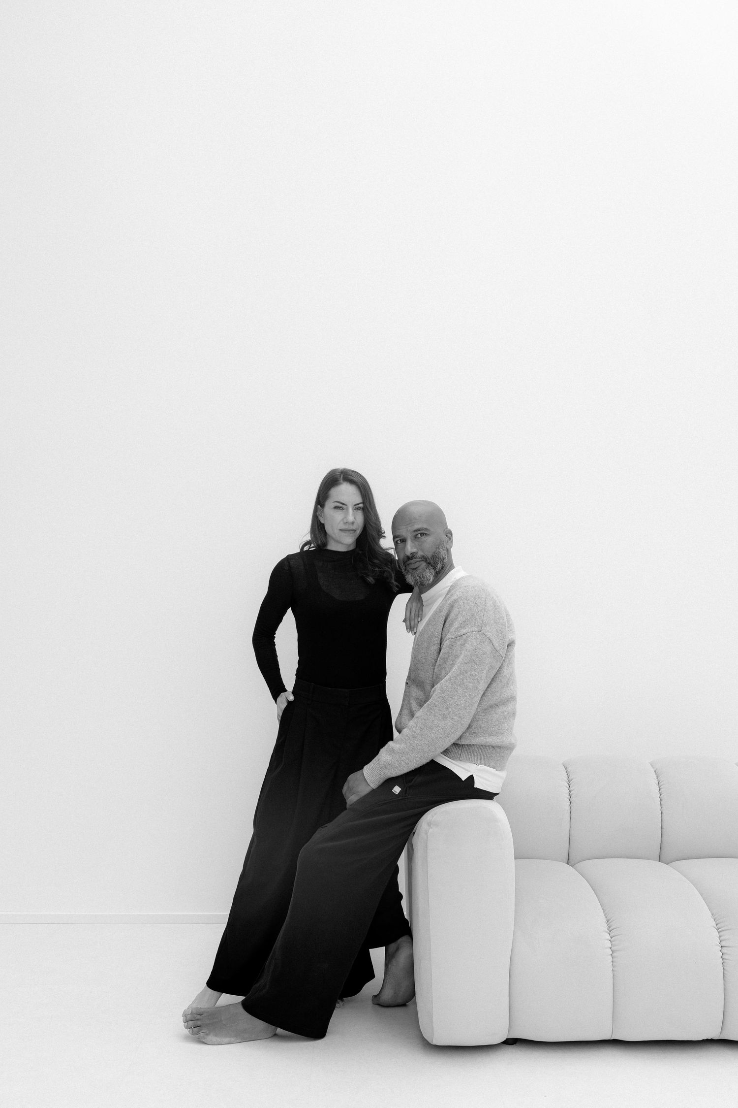 Black and white photo of a man and woman posing together near a white sofa against a bright white wall.