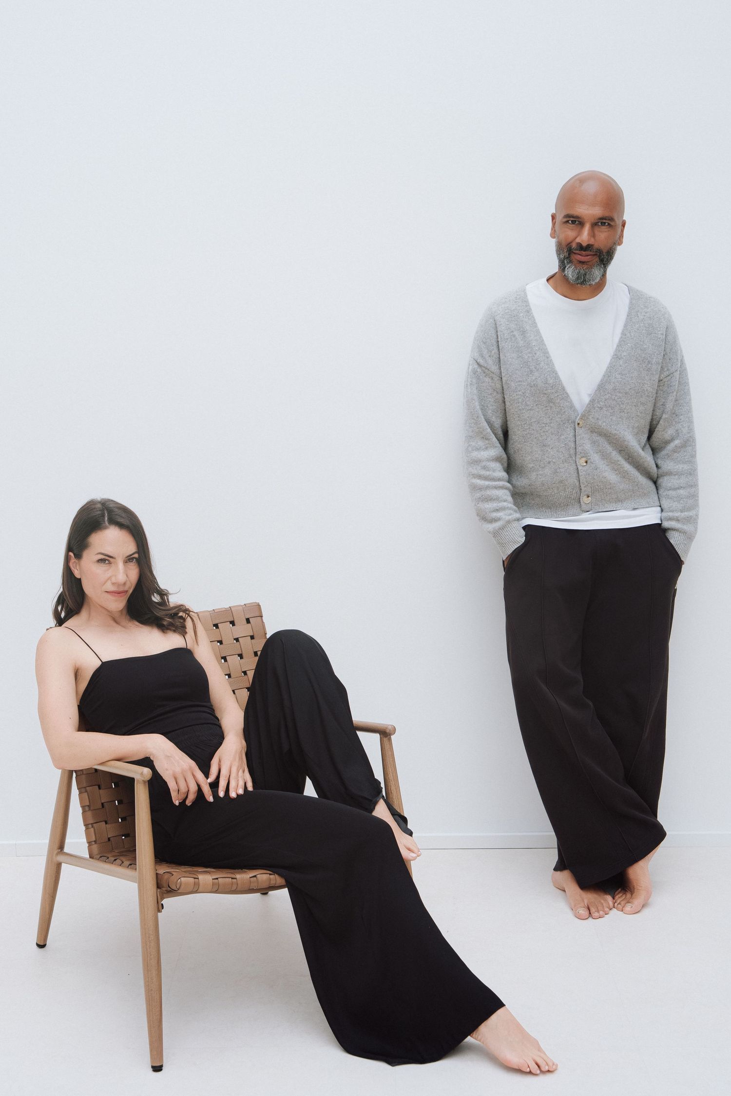A woman in black jumpsuit sits in a woven chair beside a bald man in grey cardigan and black trousers against white wall.