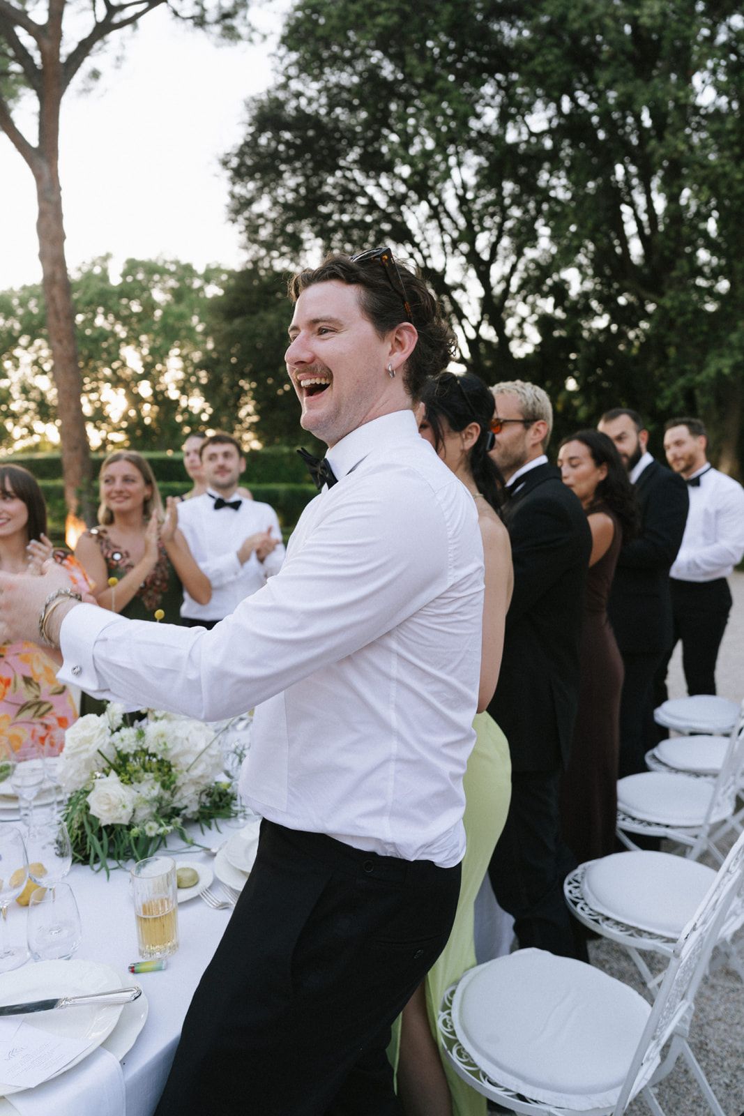 Laughing wedding guest in white shirt and black bow tie clapping at an elegant outdoor reception.