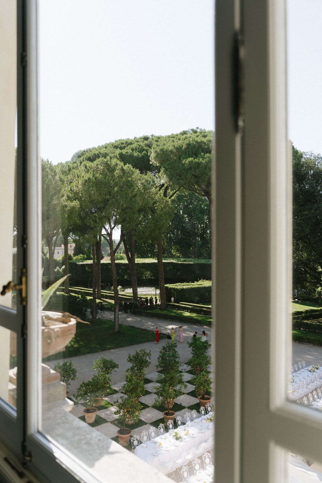 View through open white window of elegant Italian garden with tall pine trees, manicured hedges, and potted topiaries on checkered path.