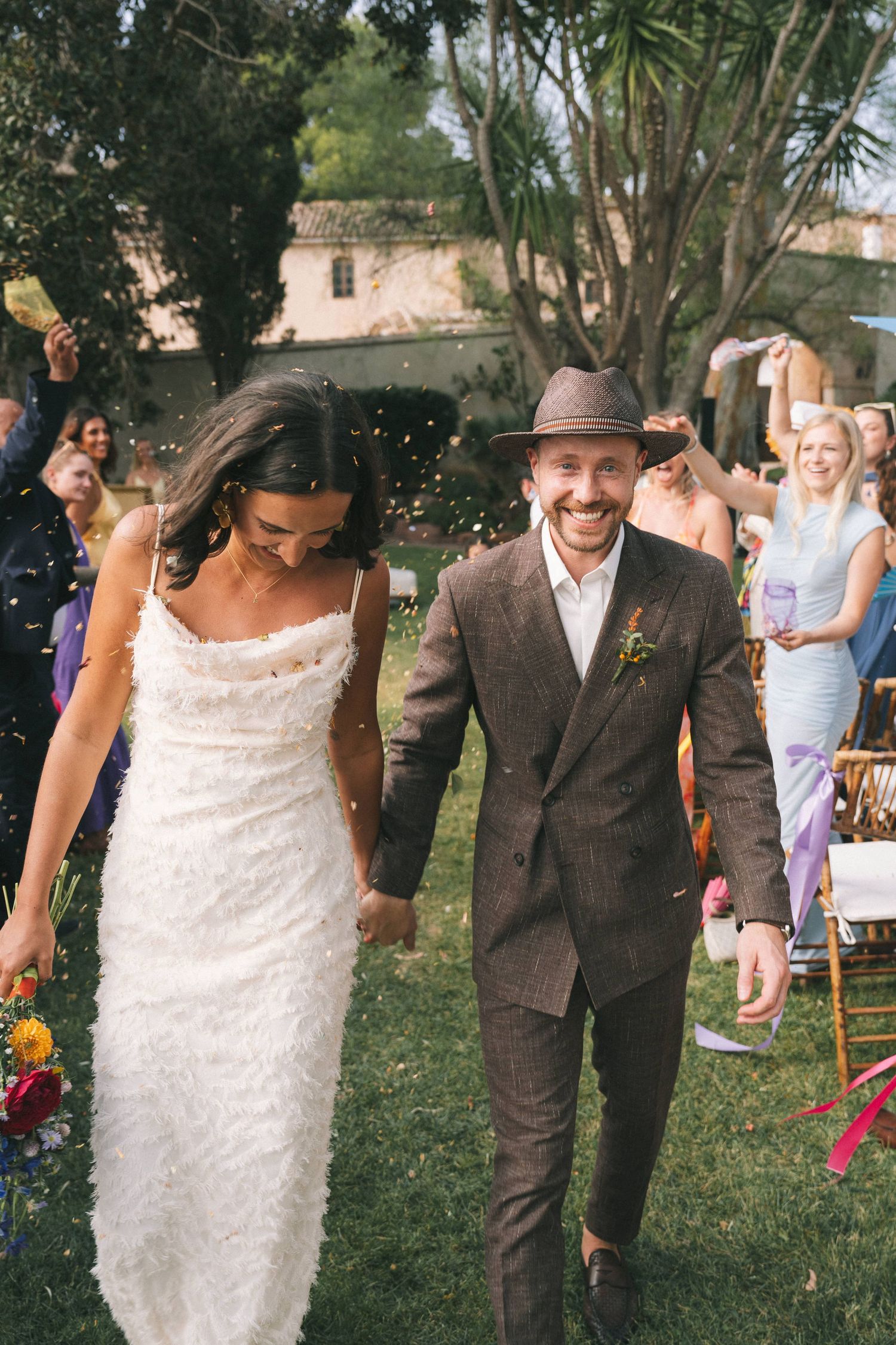 Joyful couple walks through garden path during wedding celebration with guests in background.