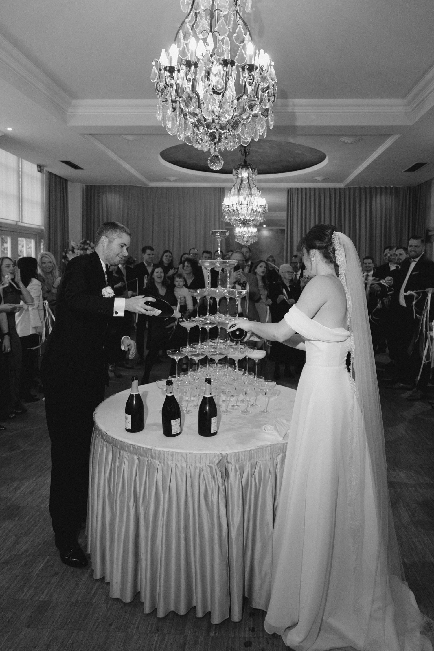 Couple pouring champagne into a tower of glasses under crystal chandeliers at their wedding reception.
