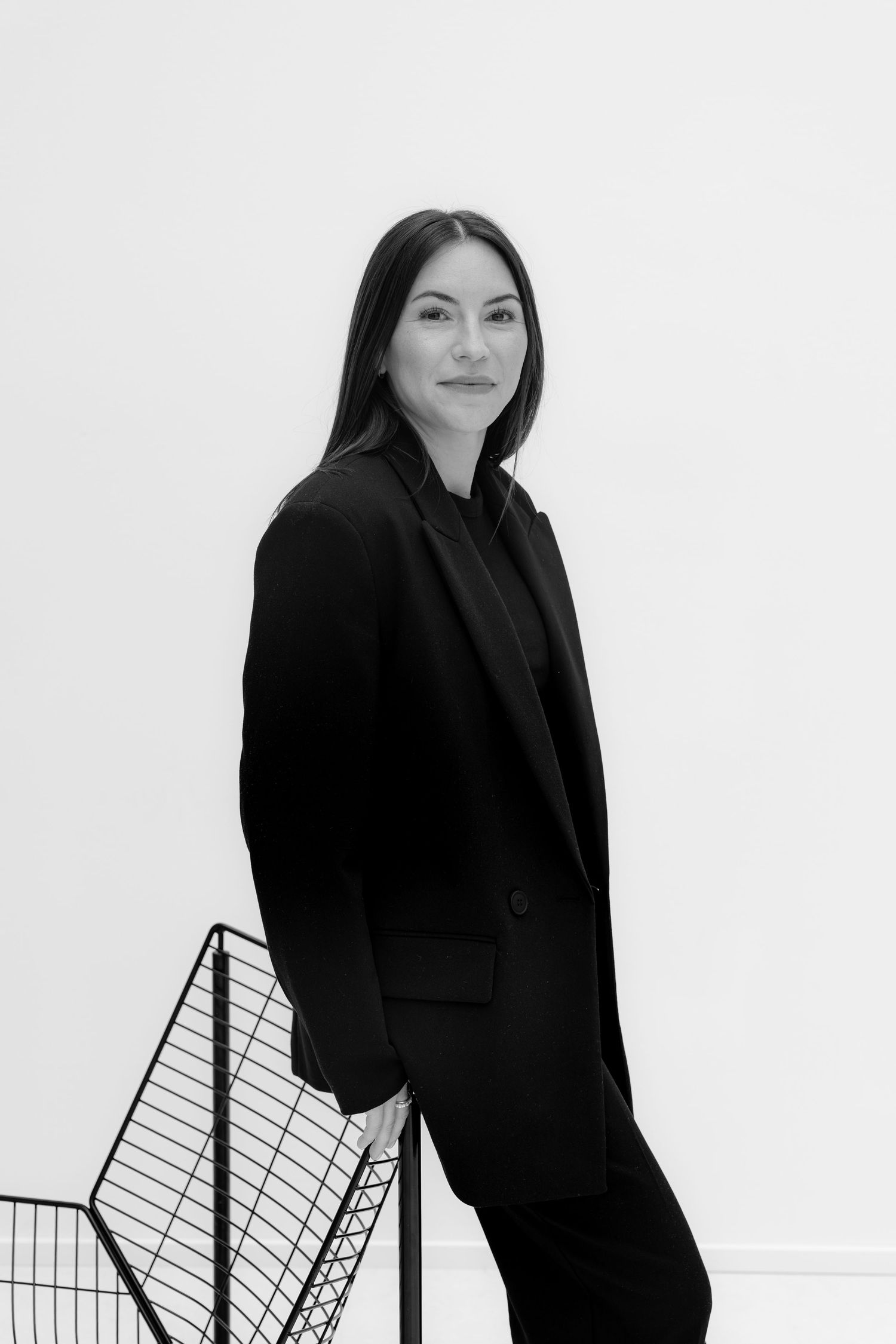 Black and white portrait of a woman in a black blazer leaning against a wire chair against a white background.