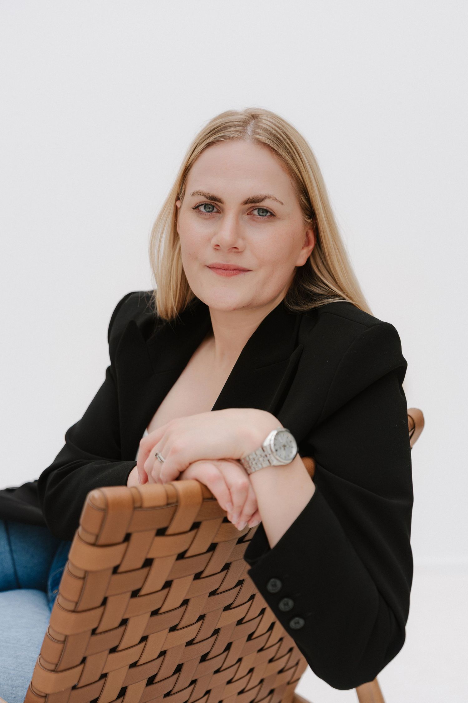 Blonde woman in black blazer sitting relaxed on woven leather chair against white background.
