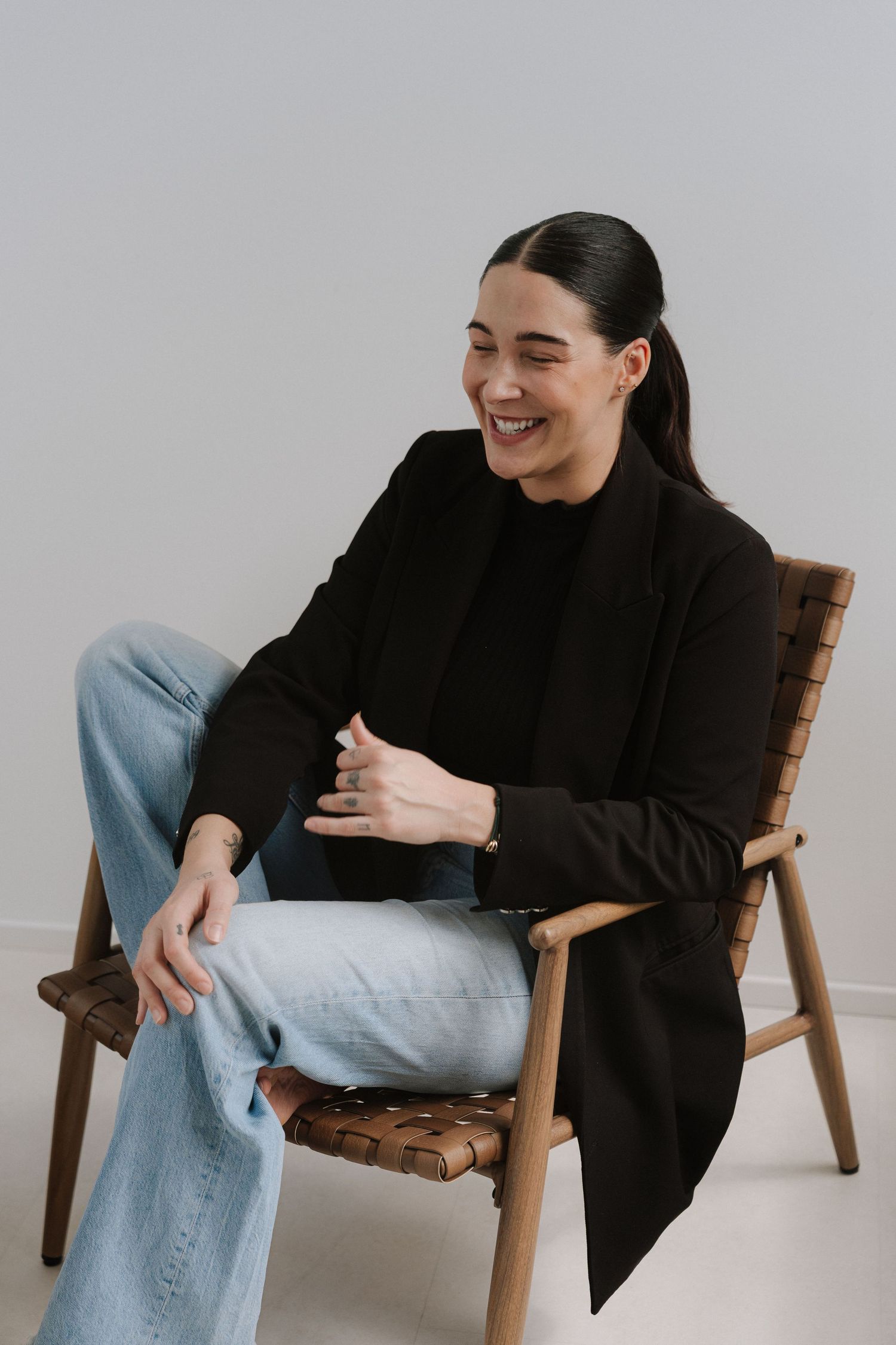 Smiling woman in black blazer and jeans sitting relaxed on a wooden leather chair against a light gray background.