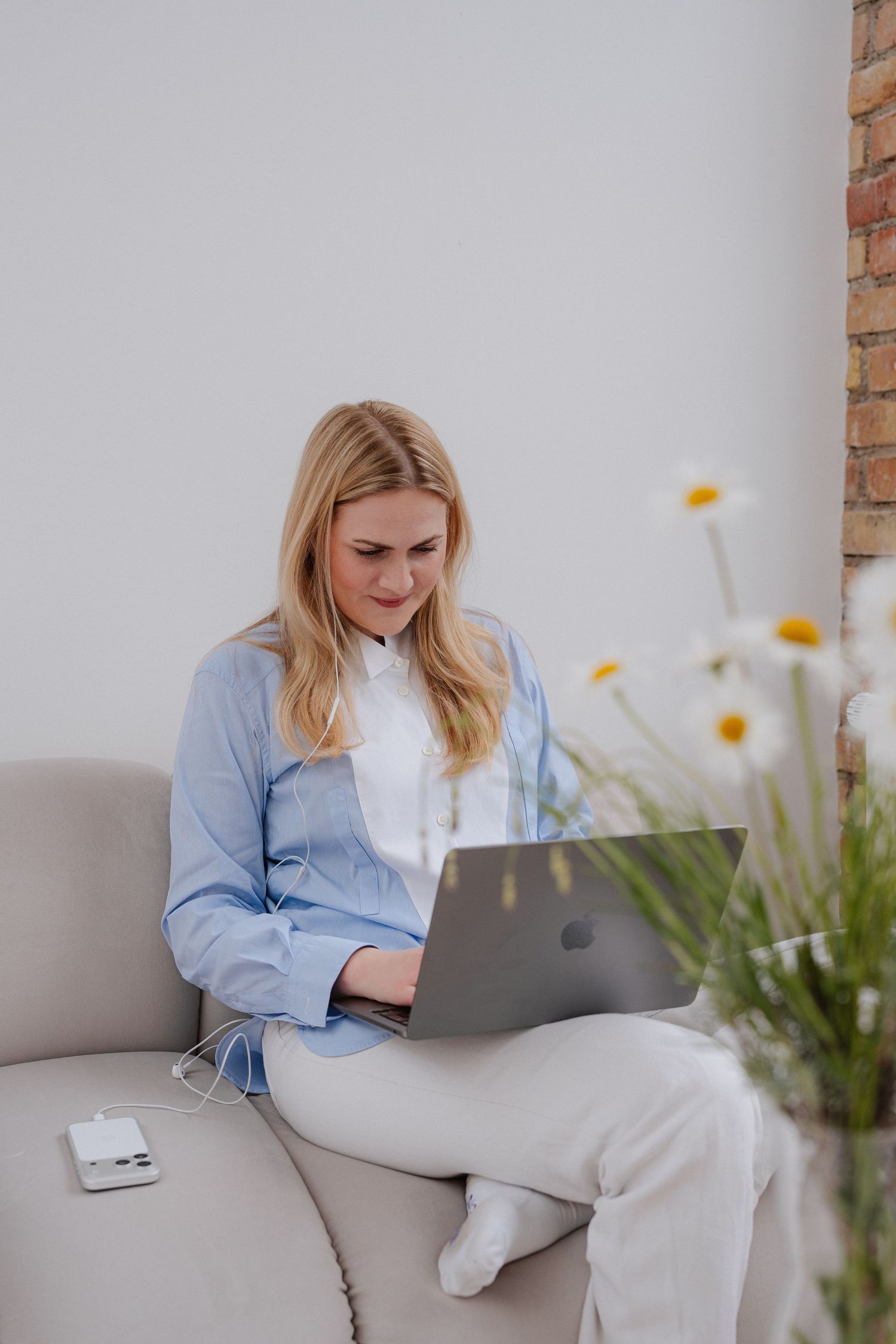 Blonde woman in blue shirt working on MacBook laptop while sitting on a beige sofa with daisies in foreground.