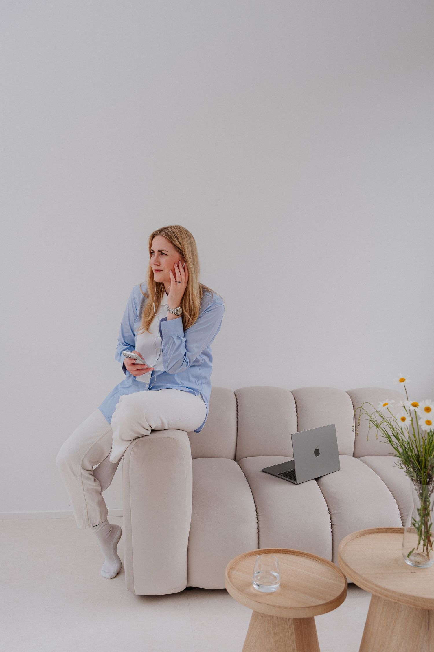 Woman in blue shirt sits on beige sofa, holding phone with thoughtful expression, laptop and flowers nearby.