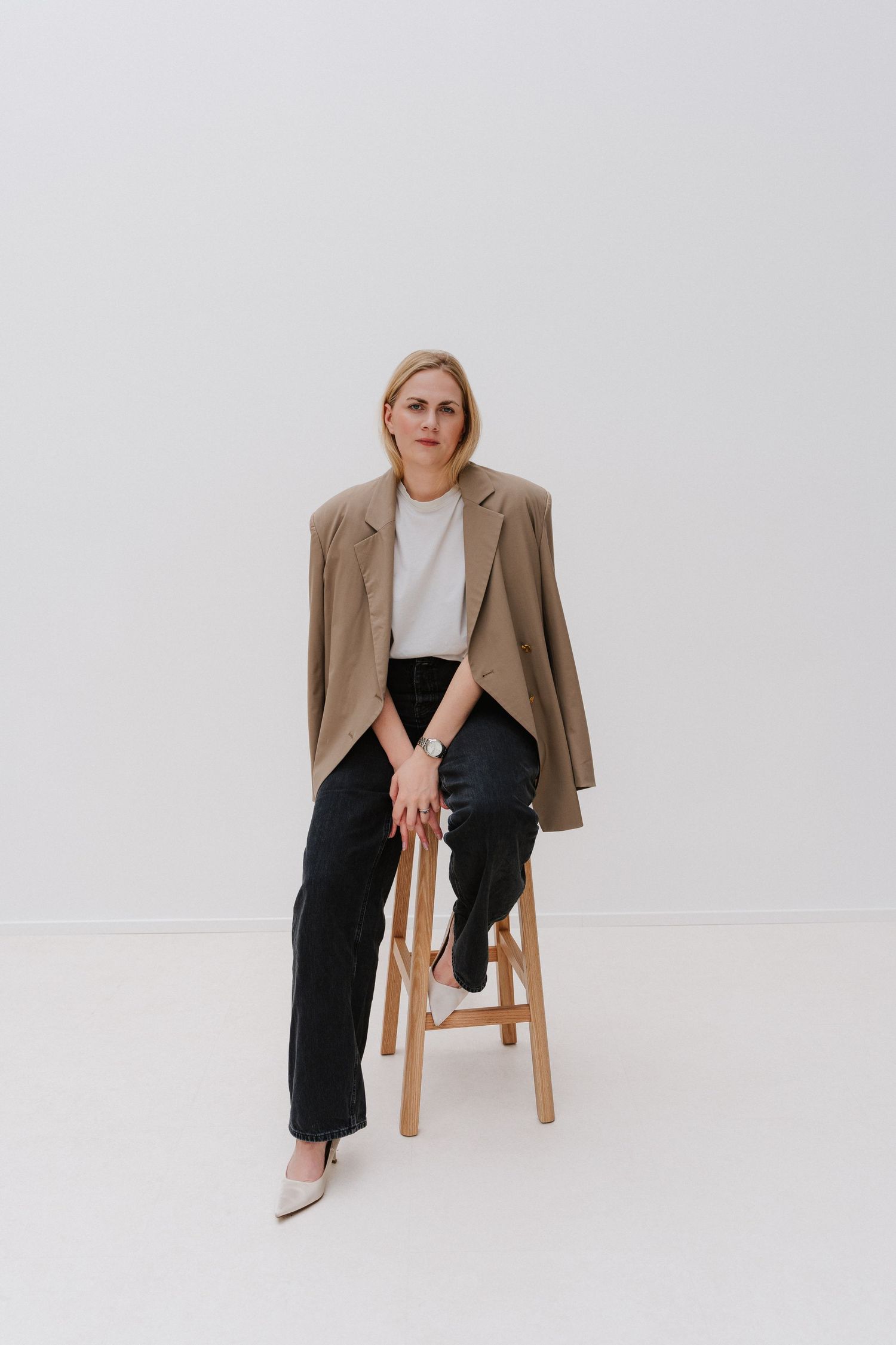 Blonde woman in beige blazer, white tee, and black pants sits on wooden stool against white background.