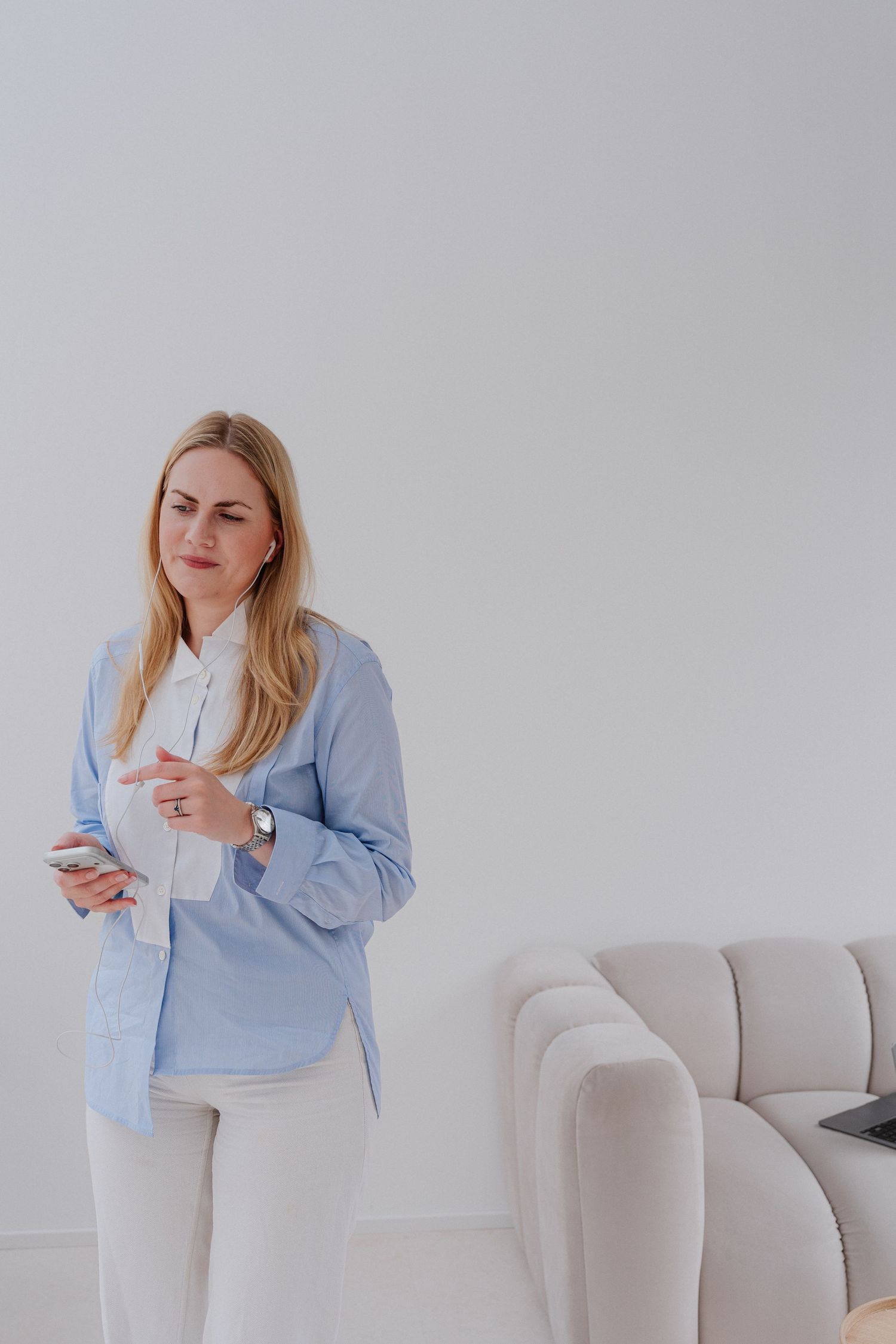 Woman in blue shirt holding smartphone standing in modern white minimalist living room with sofa.