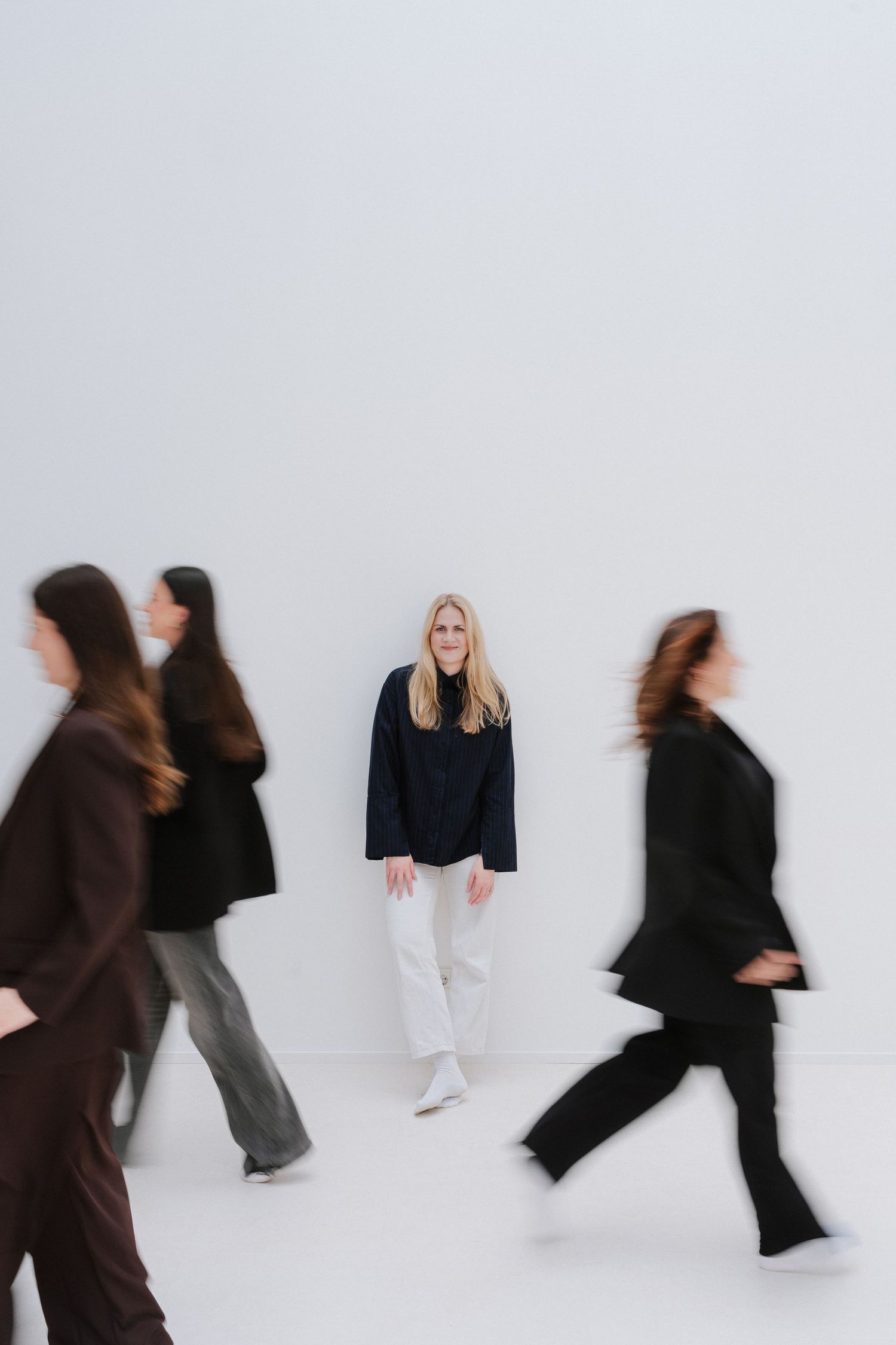 A smiling blonde woman stands still in white pants and dark jacket while blurred figures walk past her.