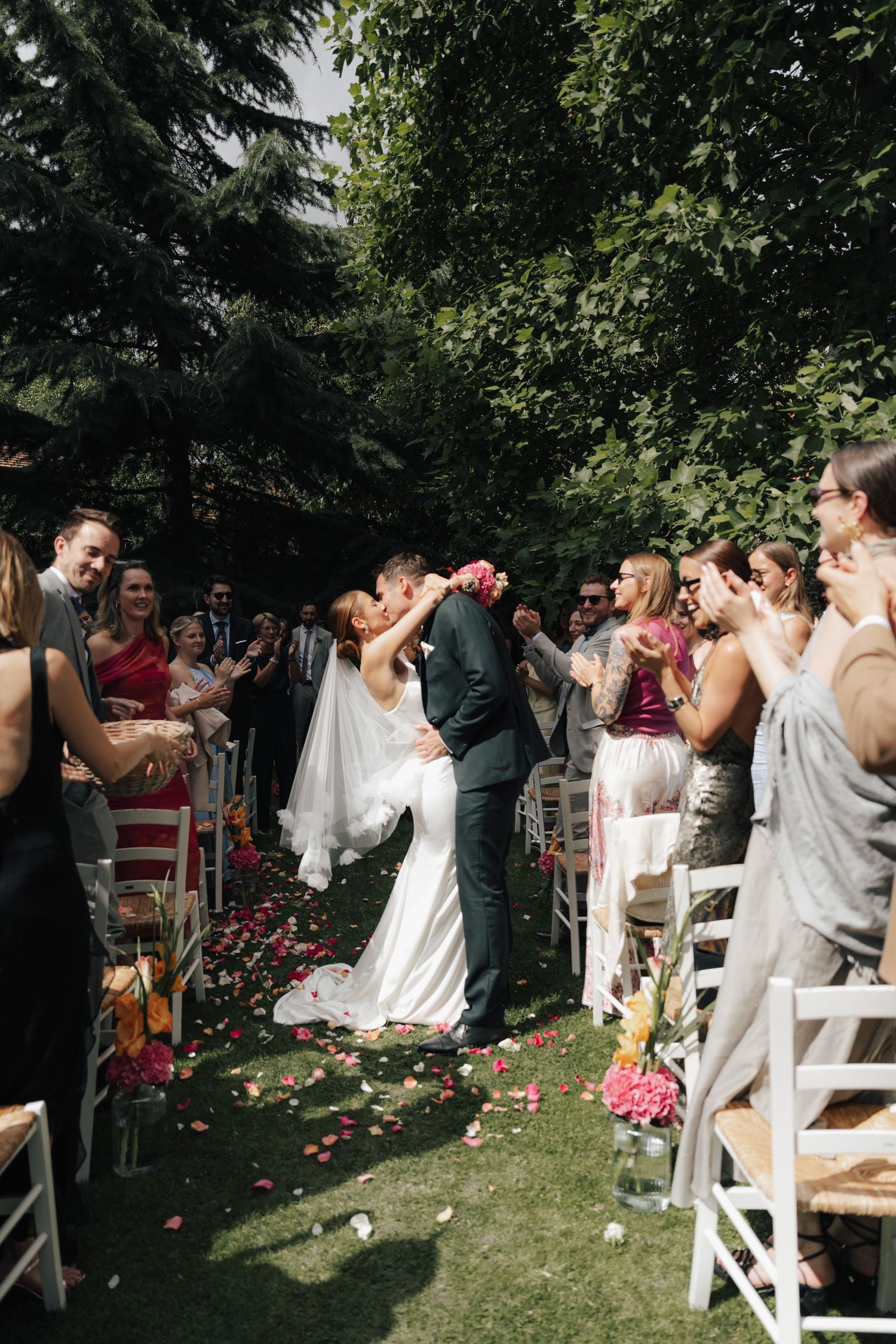 Wedding guests celebrate as couple walks down the aisle amid flower petals in a garden setting.
