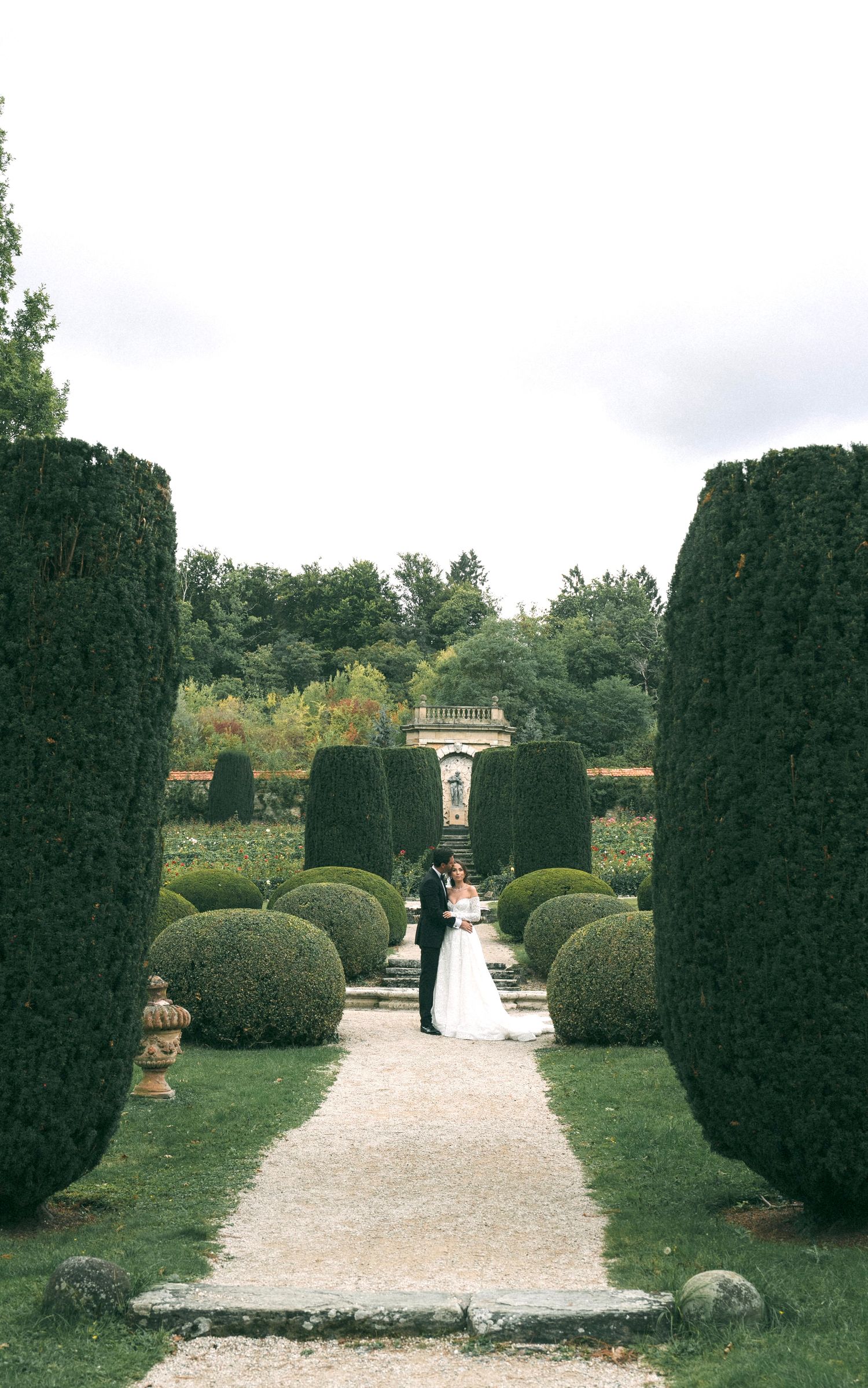 Bride and groom share a romantic moment on a garden path lined with manicured hedges and topiaries at an elegant estate.