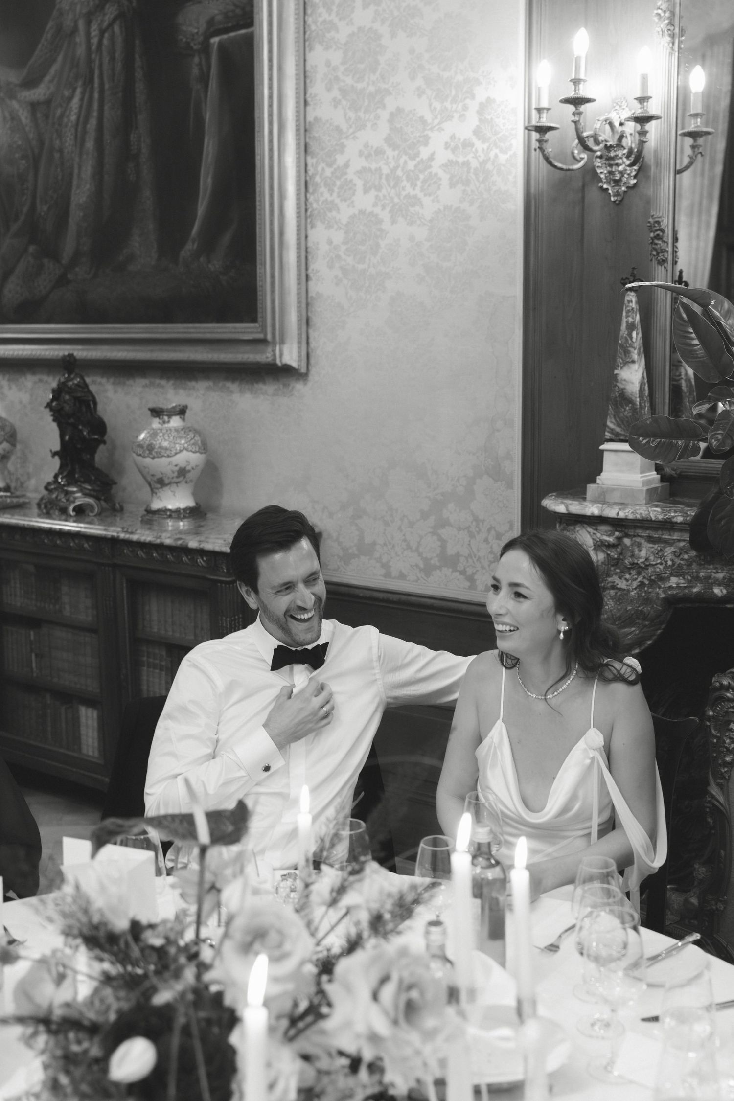 Bride and groom laugh together at candlelit reception table in elegant room, black and white wedding photo.