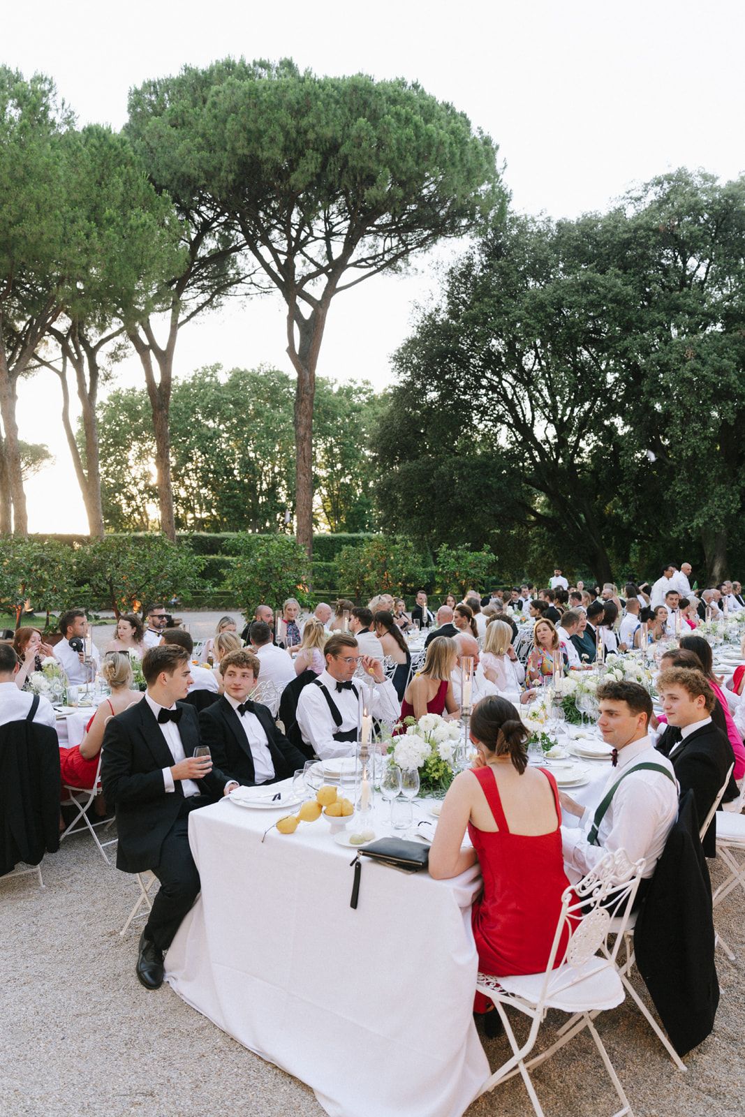 Elegant outdoor wedding reception with guests in formal attire seated at white-clothed tables surrounded by lush green trees.