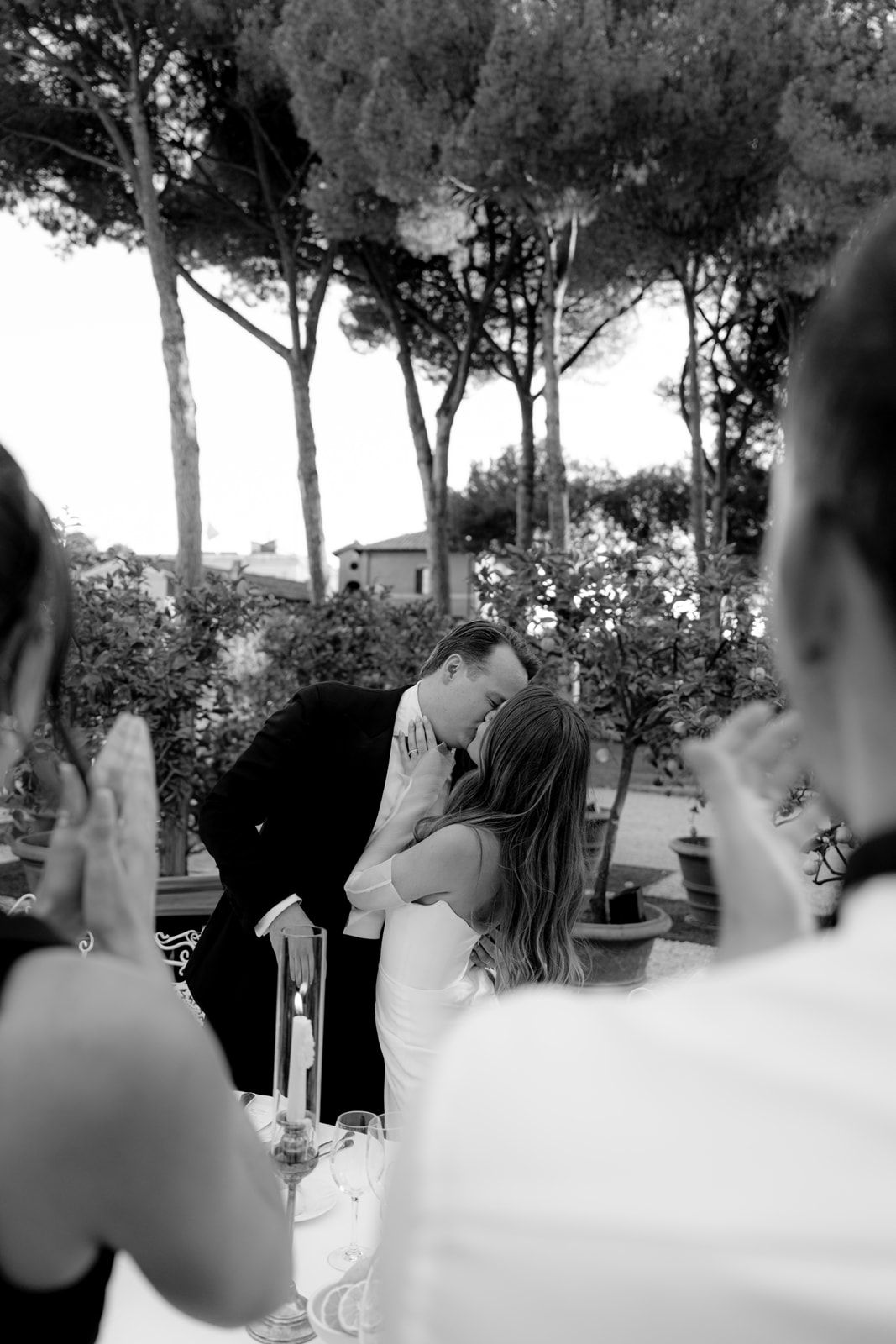 Black and white photo of a couple sharing a kiss at an outdoor wedding reception while guests applaud.