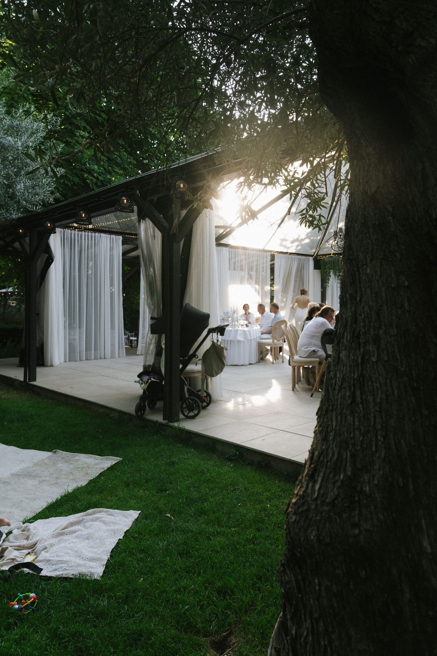 Elegant outdoor garden dining event under a white draped pergola with guests seated at tables, surrounded by lush green trees.