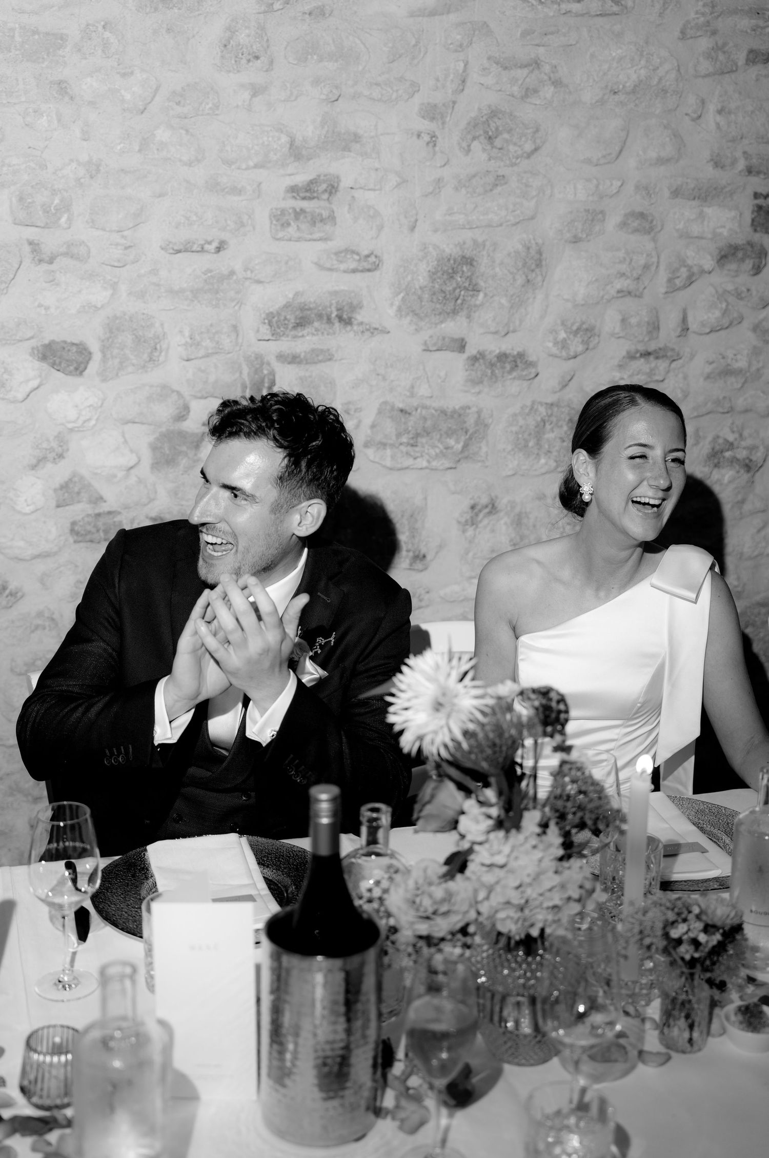 Wedding guests sharing laughter at a reception table with stone wall backdrop.