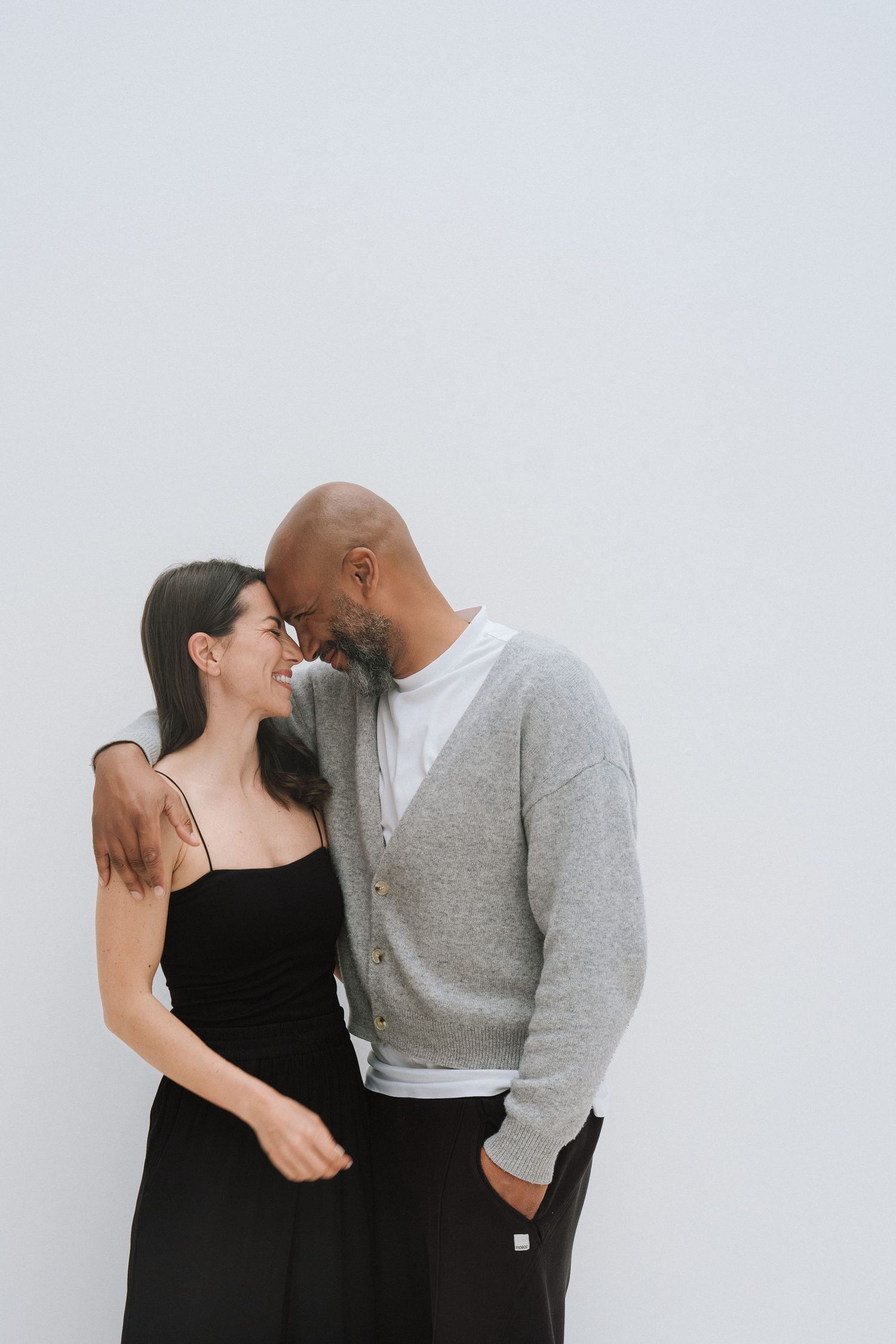 A happy interracial couple smiling and embracing against a white wall, woman in black dress, man in gray cardigan.