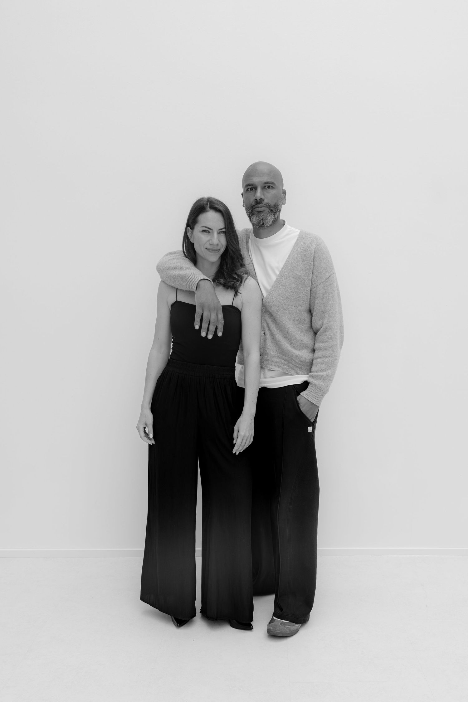 Black and white photo of a man and woman posing together against a white wall, wearing stylish dark outfits.