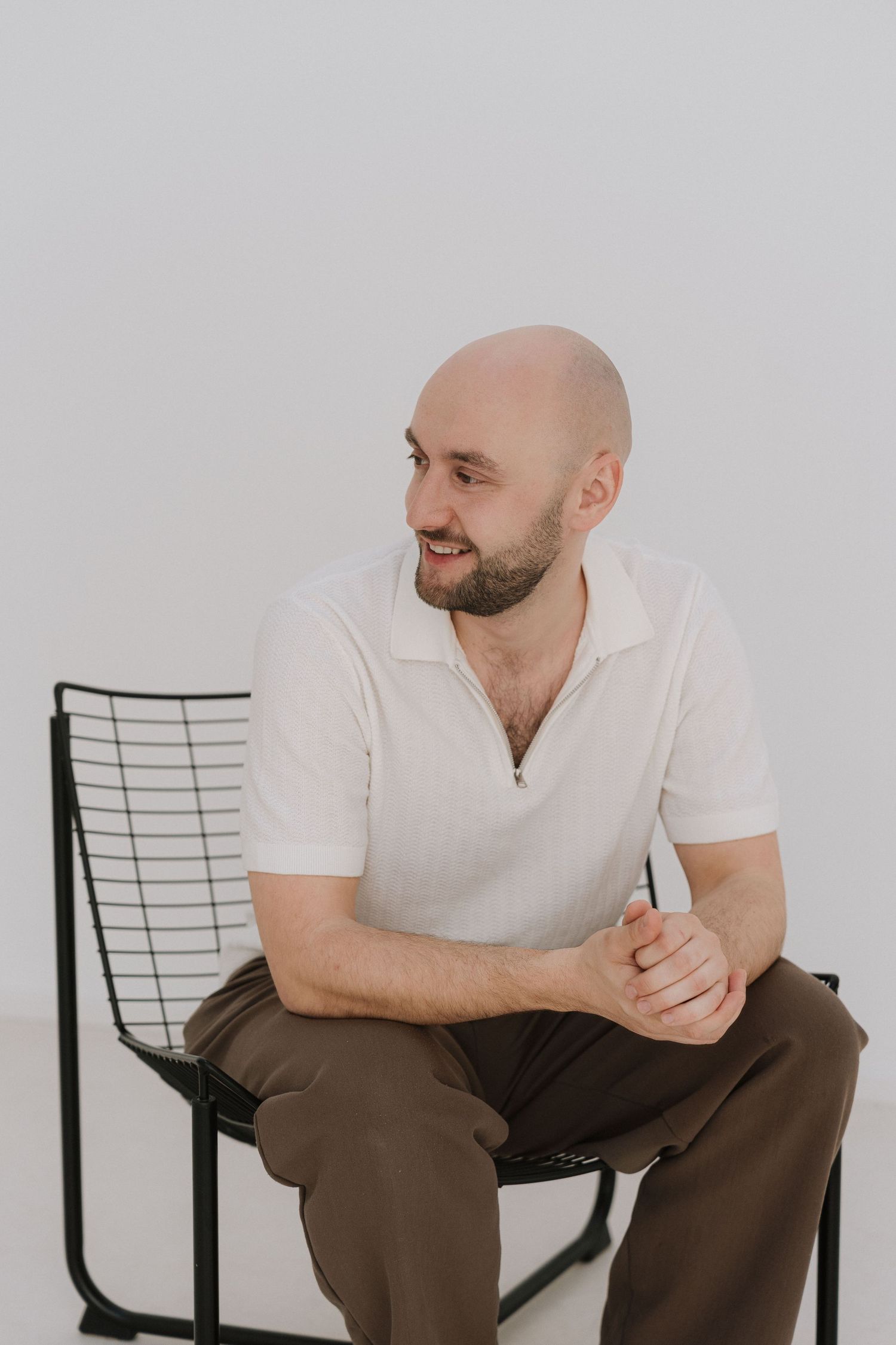 Bald bearded man in white polo shirt and brown trousers sitting on black wire chair against light gray background.