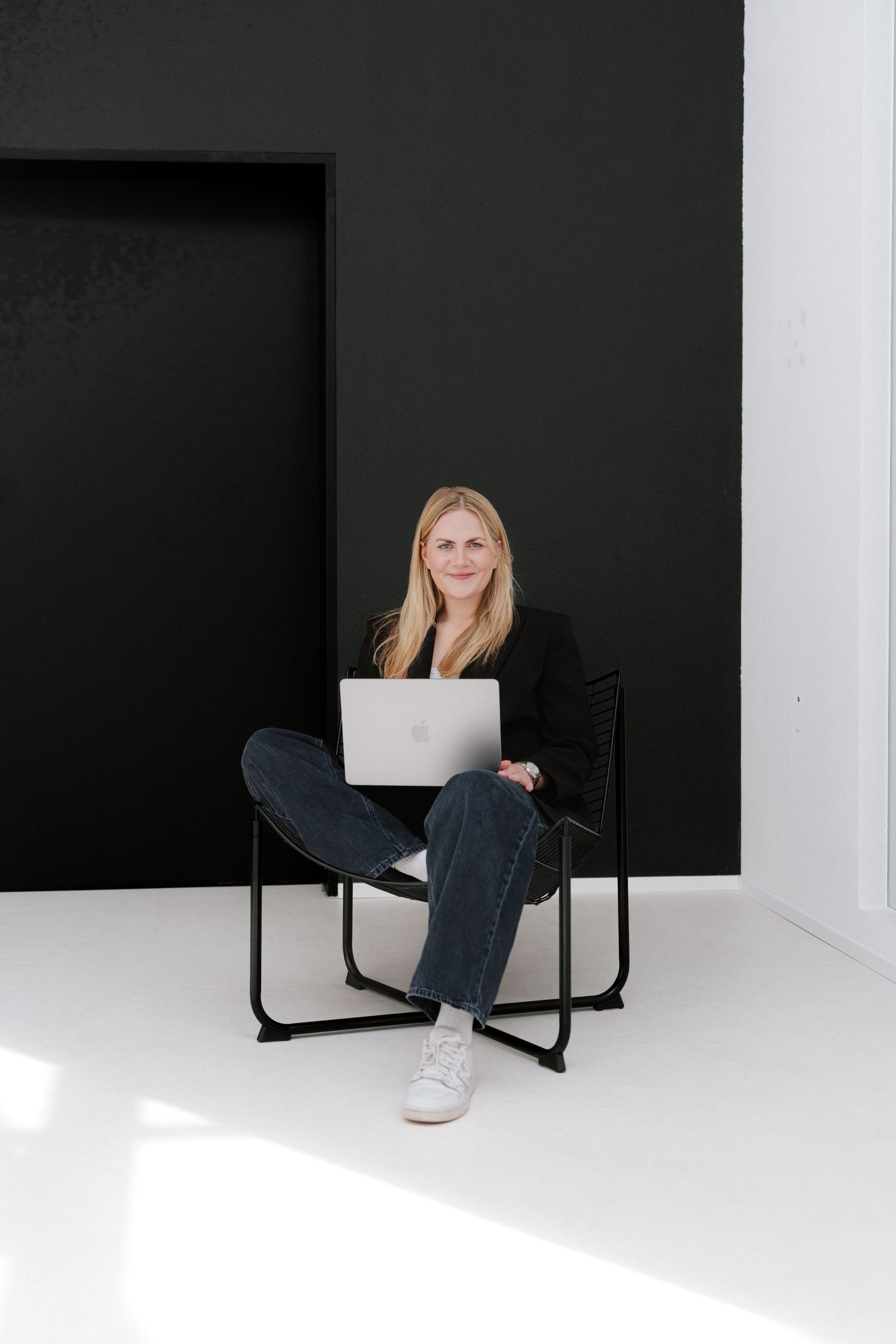 Woman sitting cross-legged on black chair with laptop, smiling against black and white studio background.