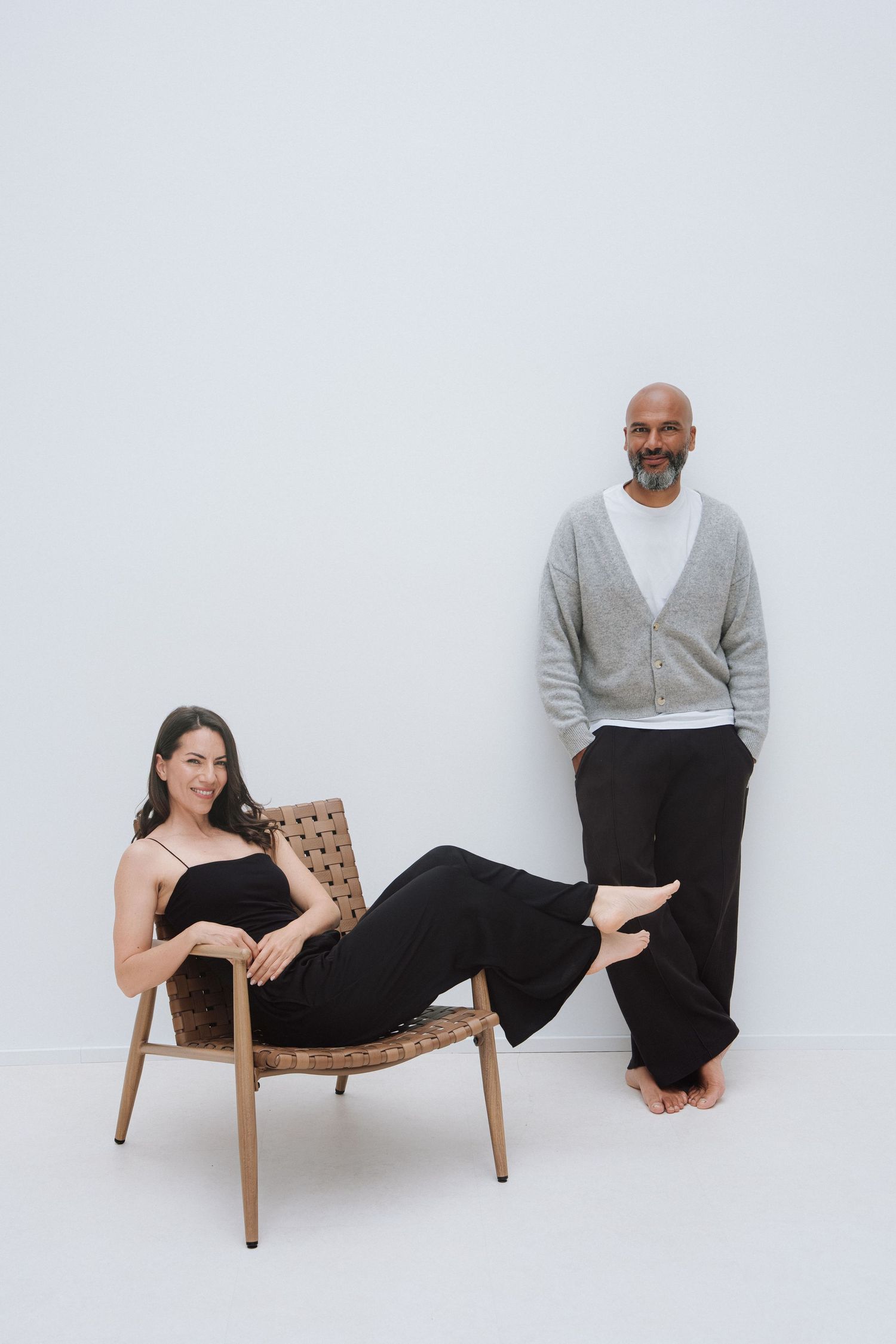 A woman in black lounges in a wooden chair beside a bald man in gray cardigan and black pants against a white wall.