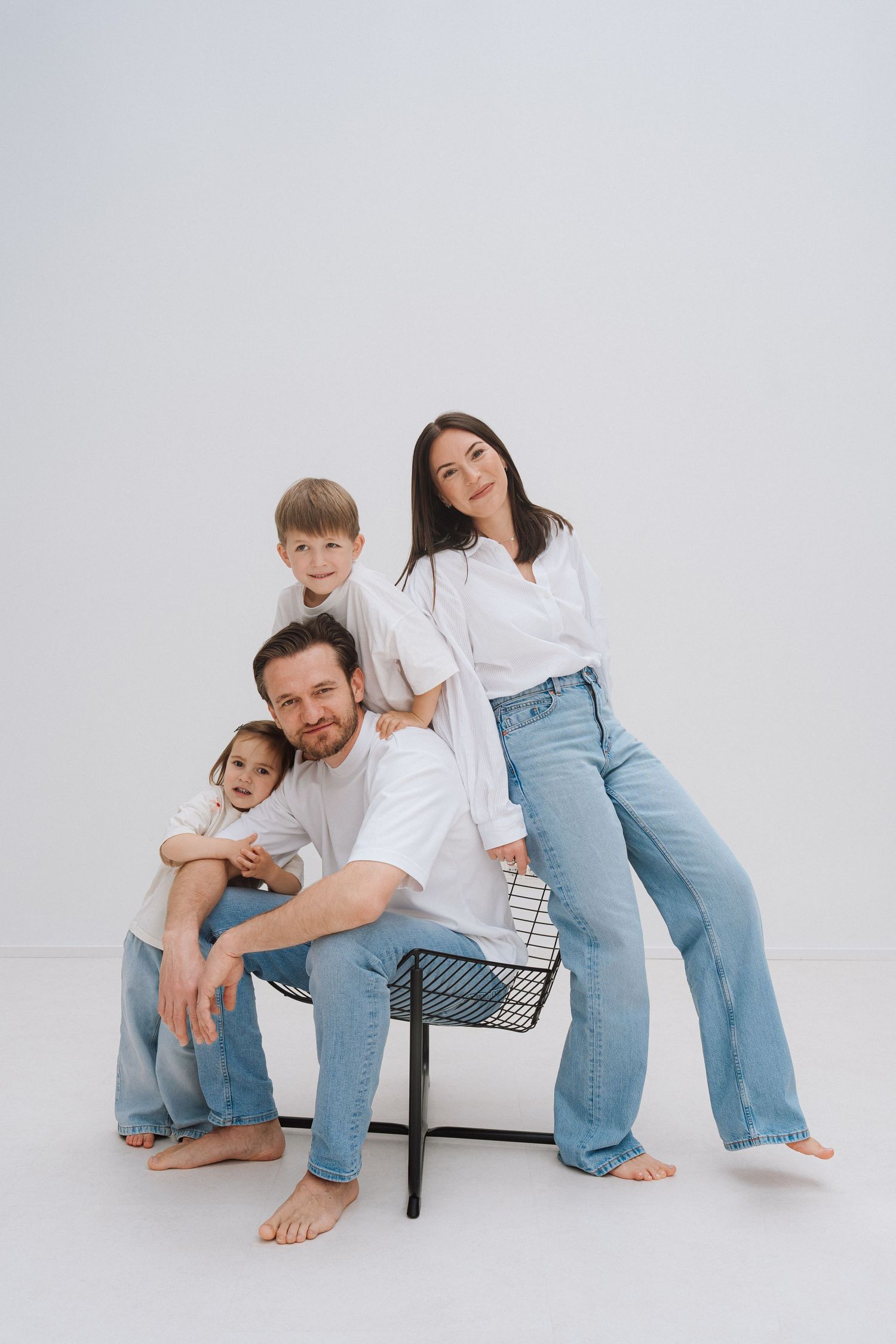 Family of four in white tops and jeans posing together in a studio, parents and two young children smiling at camera.