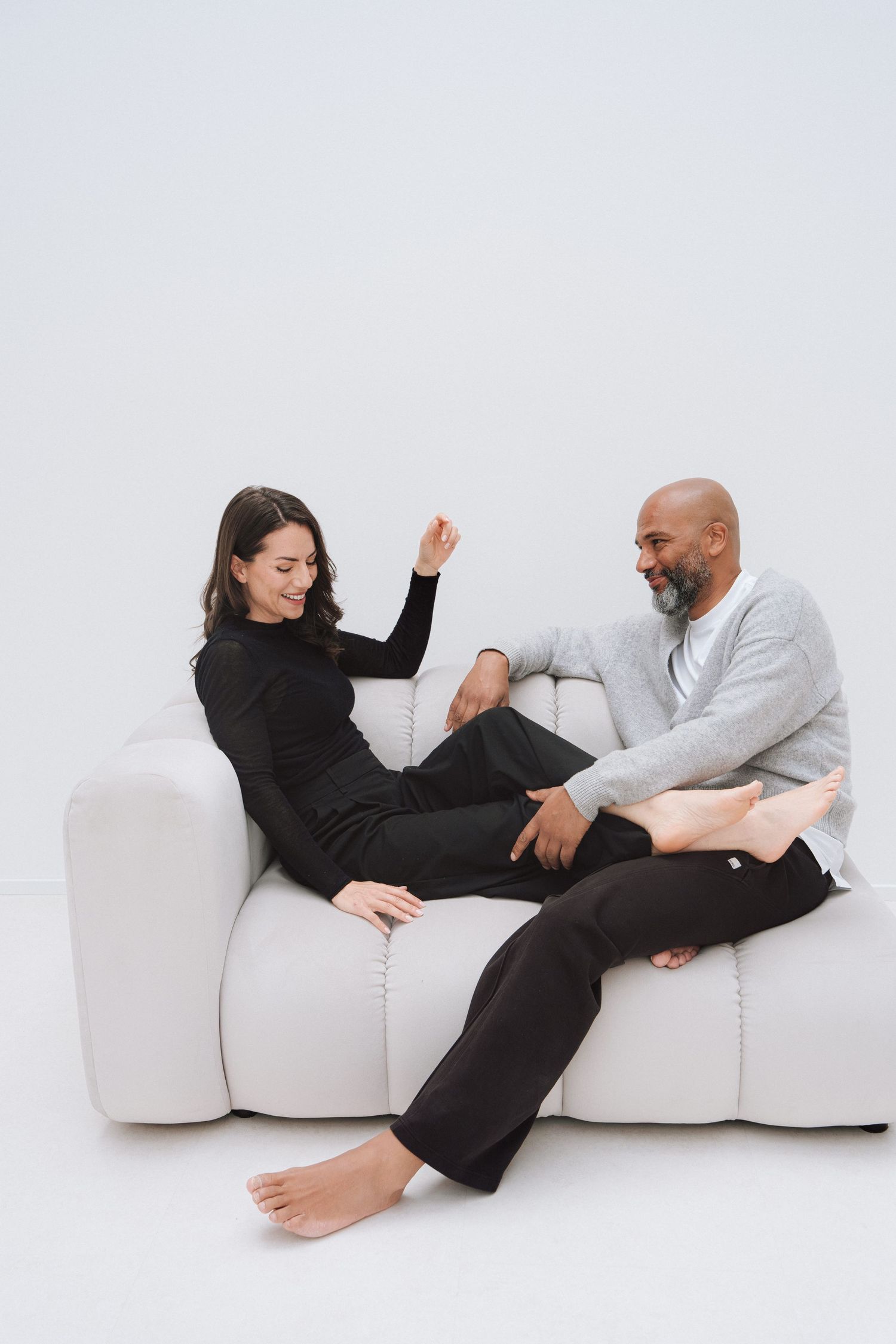 Couple relaxing on a white sofa, smiling and chatting in casual black and gray outfits against a light background.