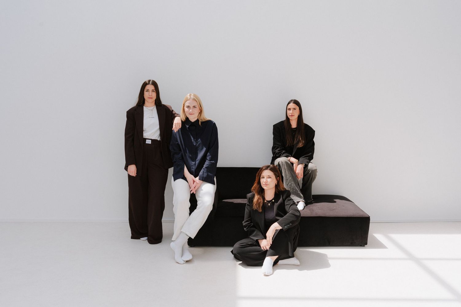 Four women in black outfits pose elegantly in a minimalist white studio setting.