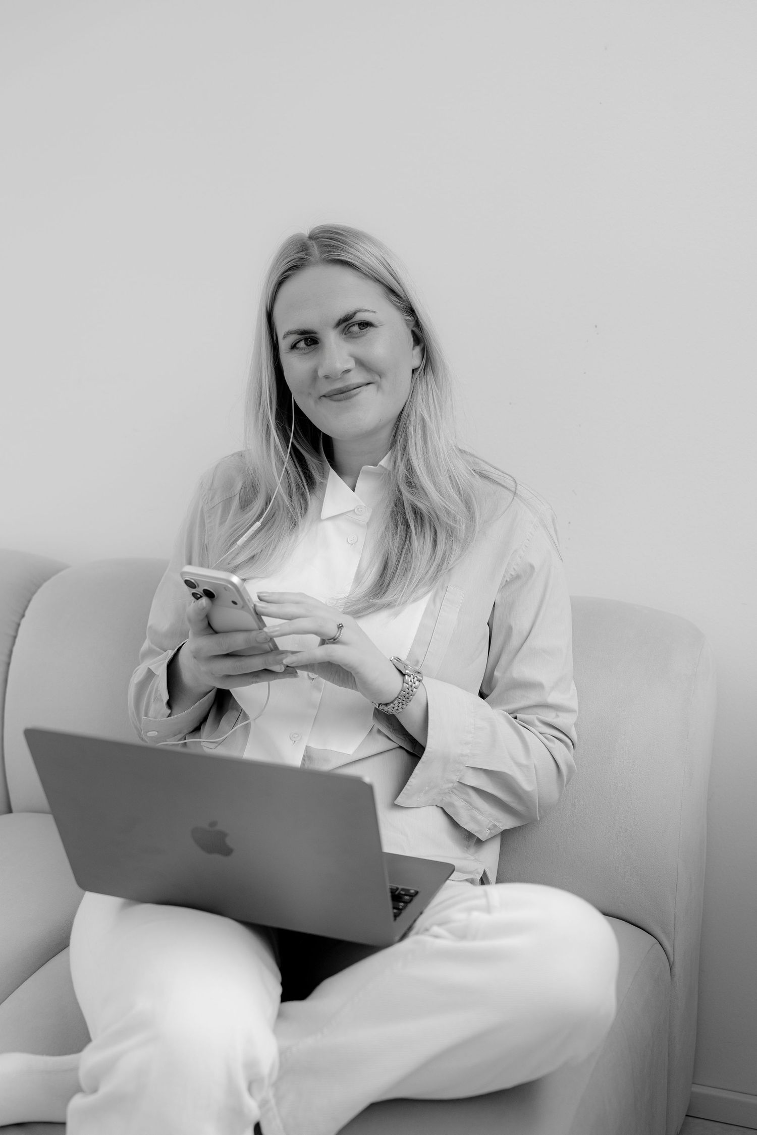 Black and white photo of a blonde woman sitting on a sofa with a laptop and smartphone, smiling at the camera.
