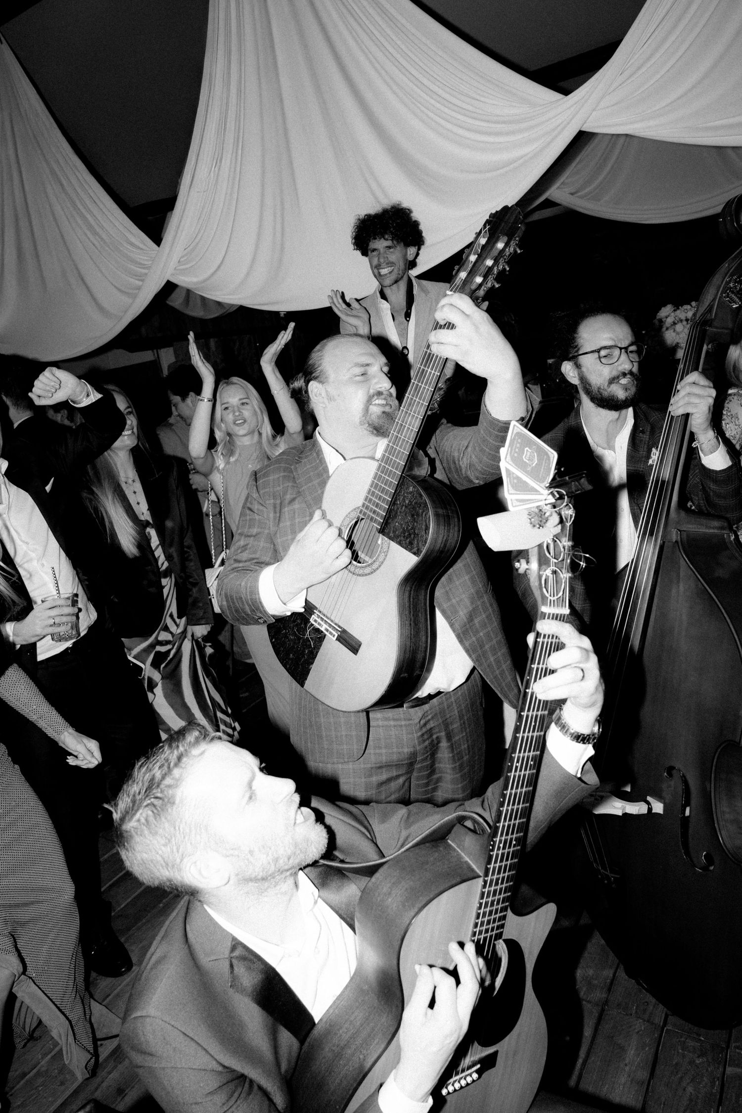 Musicians playing guitars and instruments at a lively wedding reception under draped fabric decor.