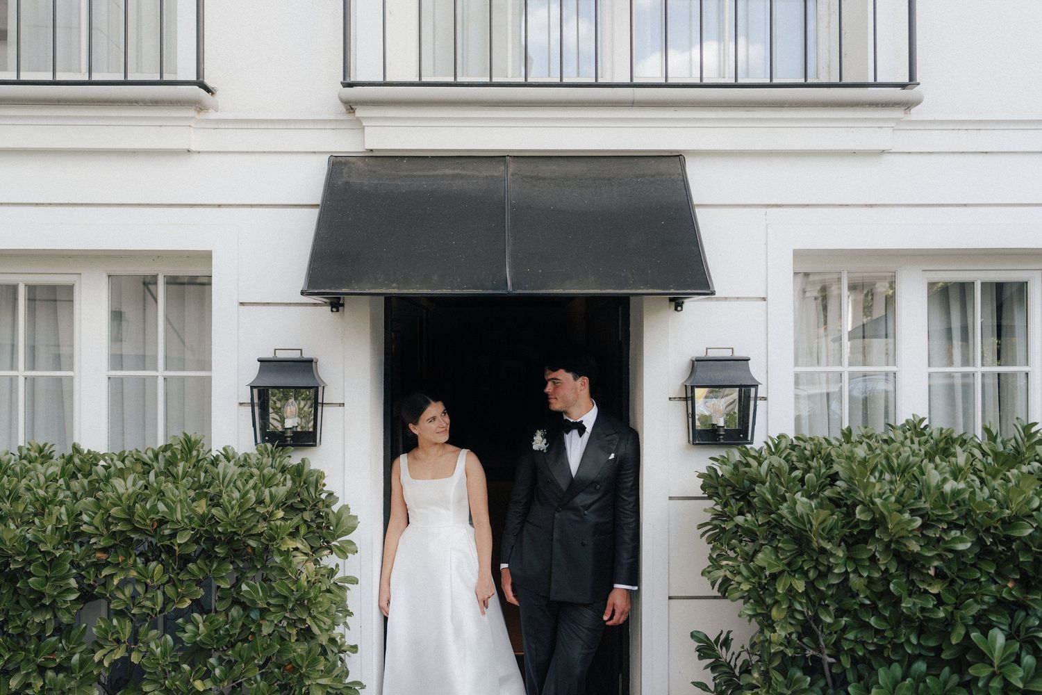 A wedding couple exits through white french doors of an elegant colonial style building.