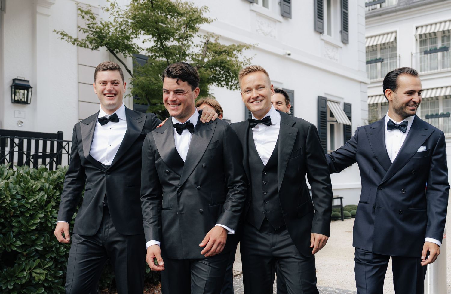 Wedding party in black tuxedos posing together outside a white building.