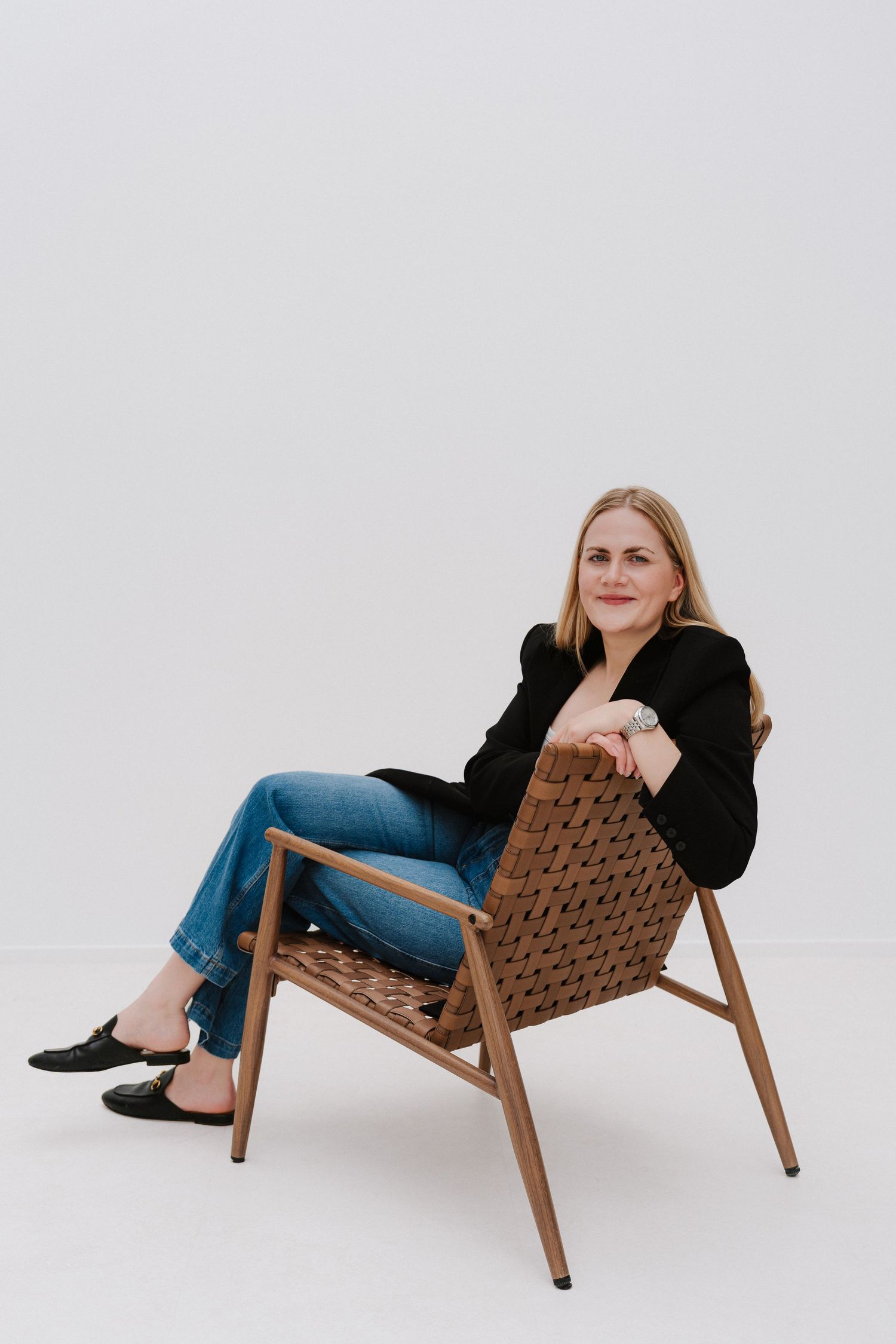 Woman in black blazer and jeans sitting relaxed in a woven wooden chair against a white background.