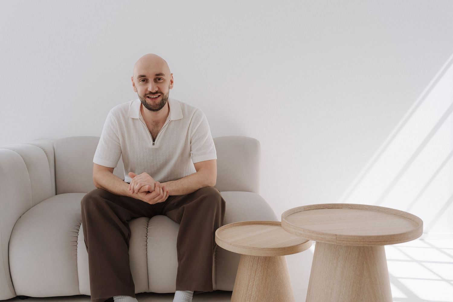 Bald man smiling, seated on a white sofa in a minimalist room with wooden side tables.
