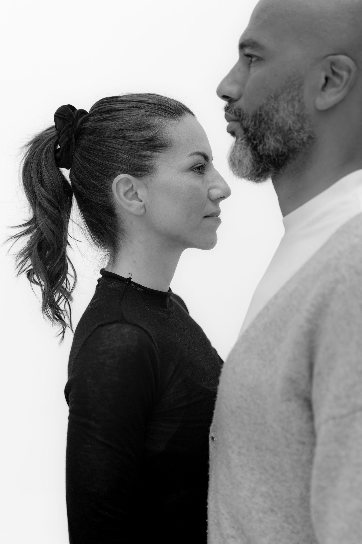 Black and white profile portrait of a woman and bearded man facing each other against a white background.