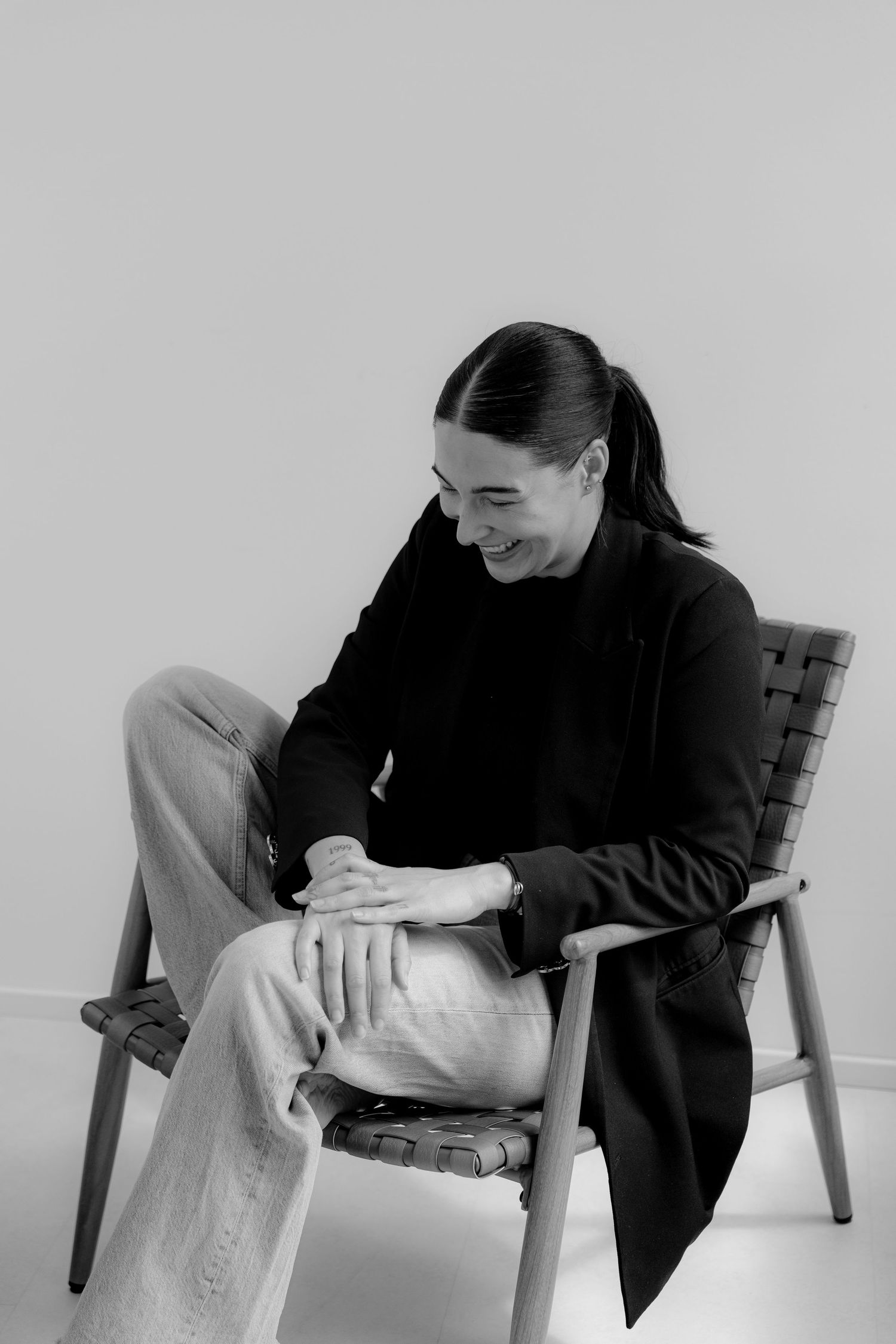 Black and white photo of a smiling woman with ponytail sitting cross-legged on a wooden chair, wearing a black blazer.